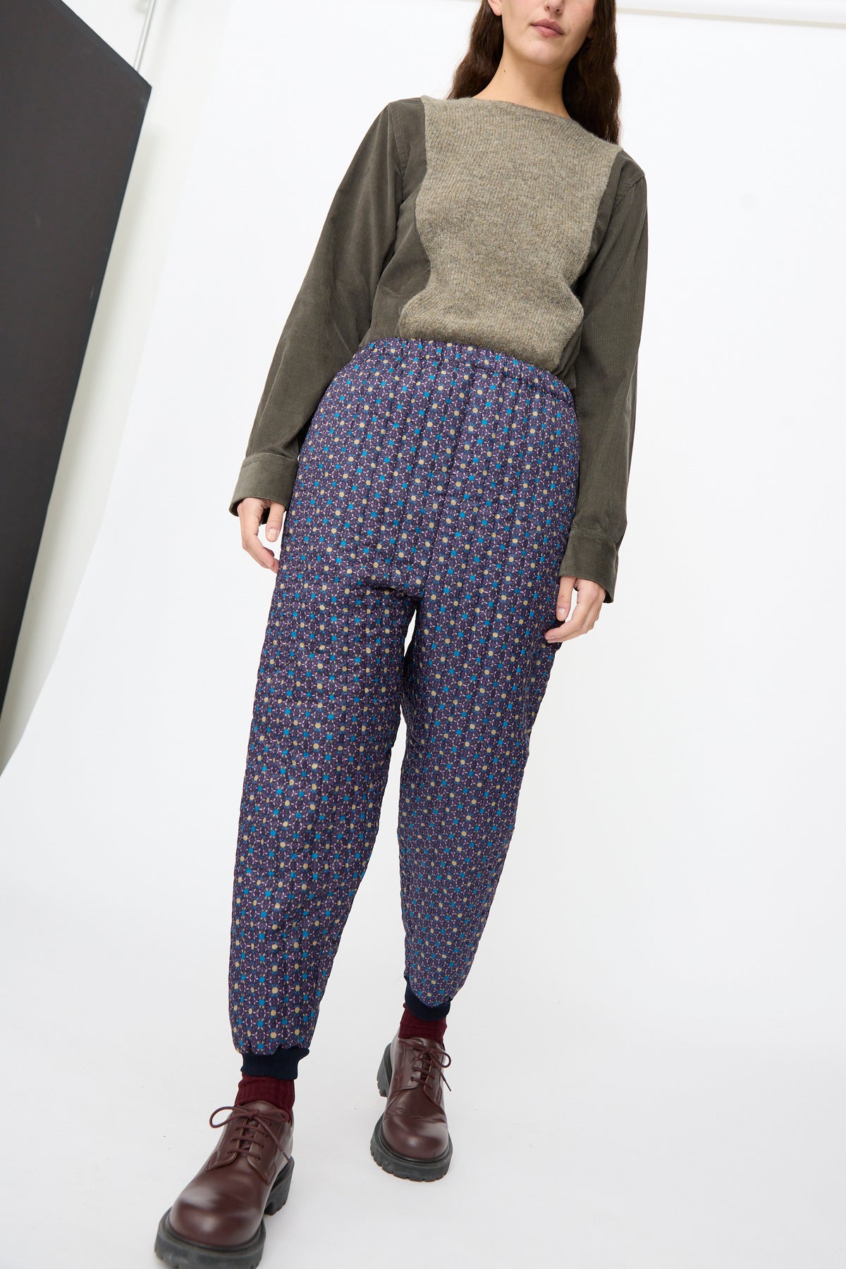 A person wearing Bless No. 08 Monpe Pants in Floral Print II, which are tapered nylon trousers with an elastic waist, paired with a two-toned knit top and dark brown lace-up boots.