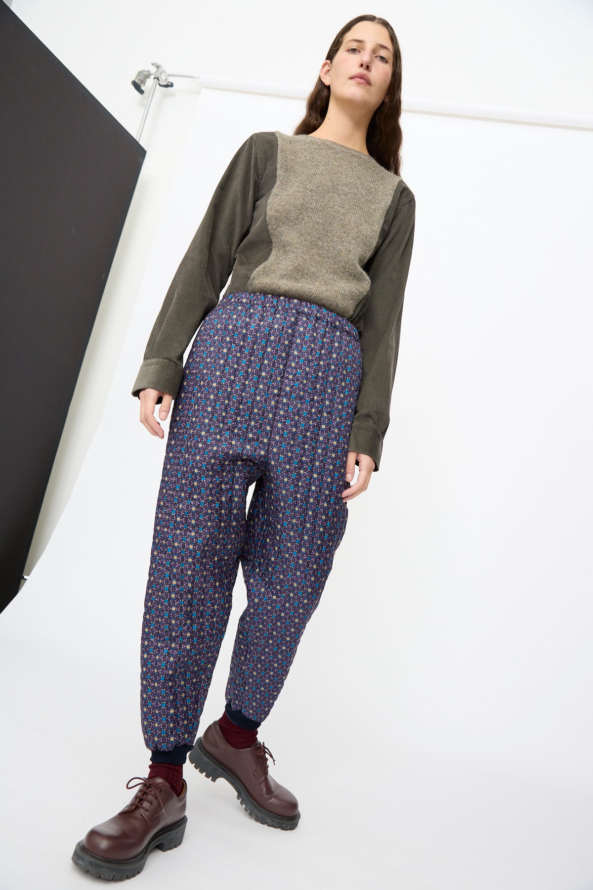 A person wearing a grey long-sleeved shirt, Bless No. 08 Monpe Pants in Floral Print II, and brown leather shoes.