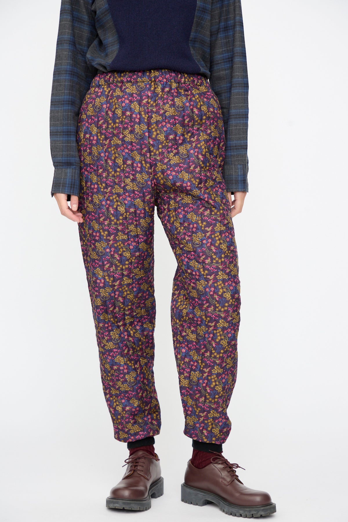 Bless No. 08 Monpe Pants in Floral Print III: loose-fitting dark purple floral print pants with an elastic waistband, worn with a dark plaid shirt and brown lace-up shoes.