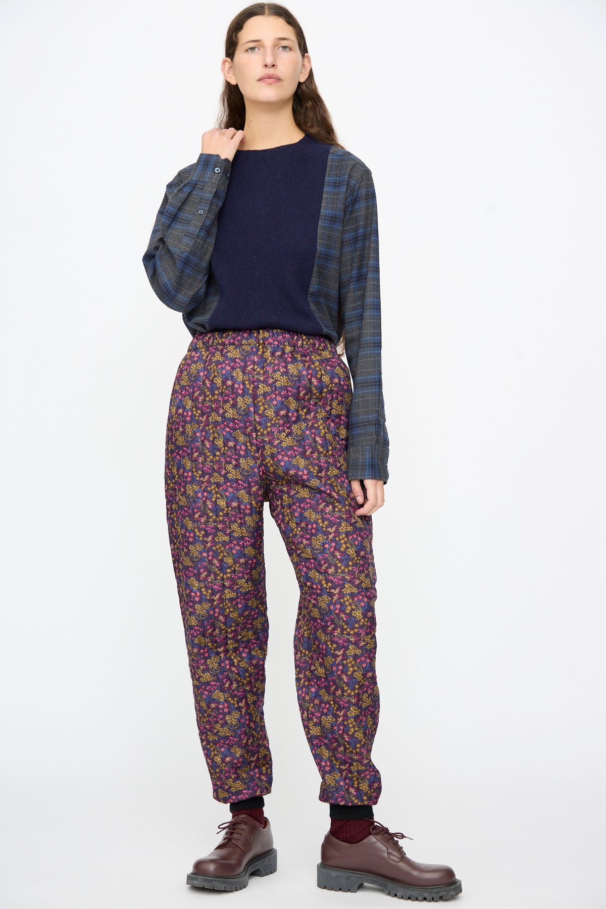 A person wearing a dark blue sweater over a plaid button-down shirt, paired with Bless No. 08 Monpe Pants in Floral Print III and dark brown lace-up shoes.