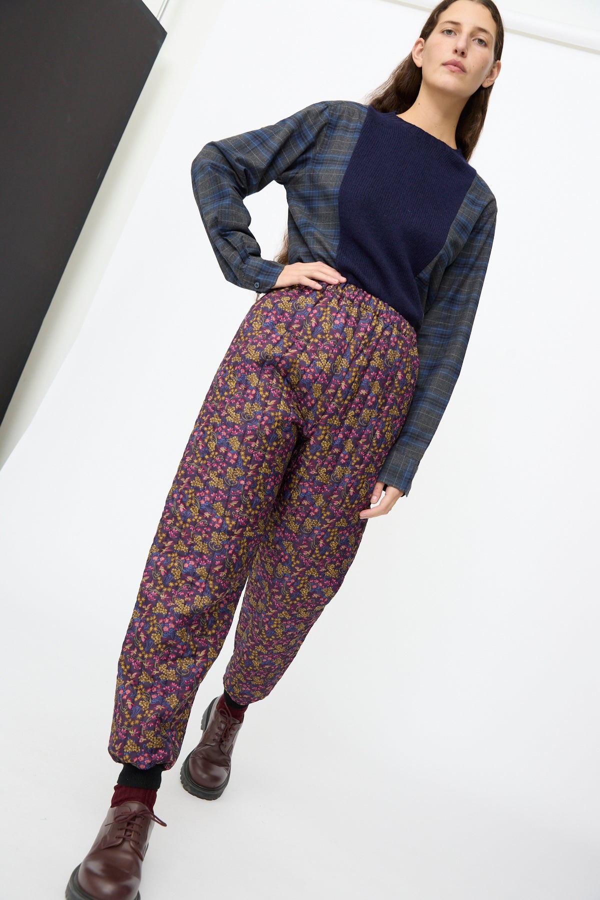 A person wearing Bless No. 08 Monpe Pants in Floral Print III, featuring a dark floral print and elastic waistband, styled with a navy blue sweater, plaid shirt, and dark leather shoes.