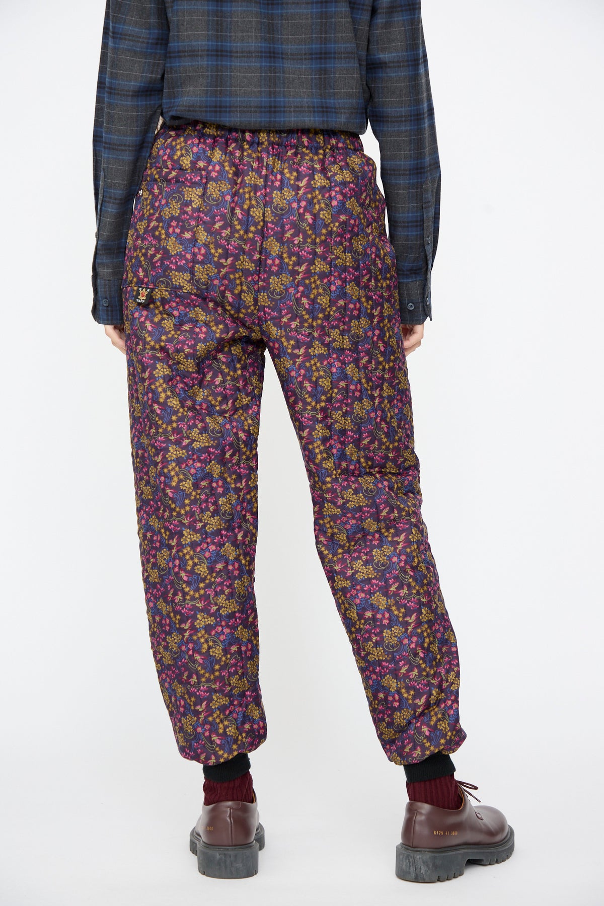 A person wearing a dark blue plaid shirt and Bless No. 08 Monpe Pants in Floral Print III, featuring a multi-colored floral print.