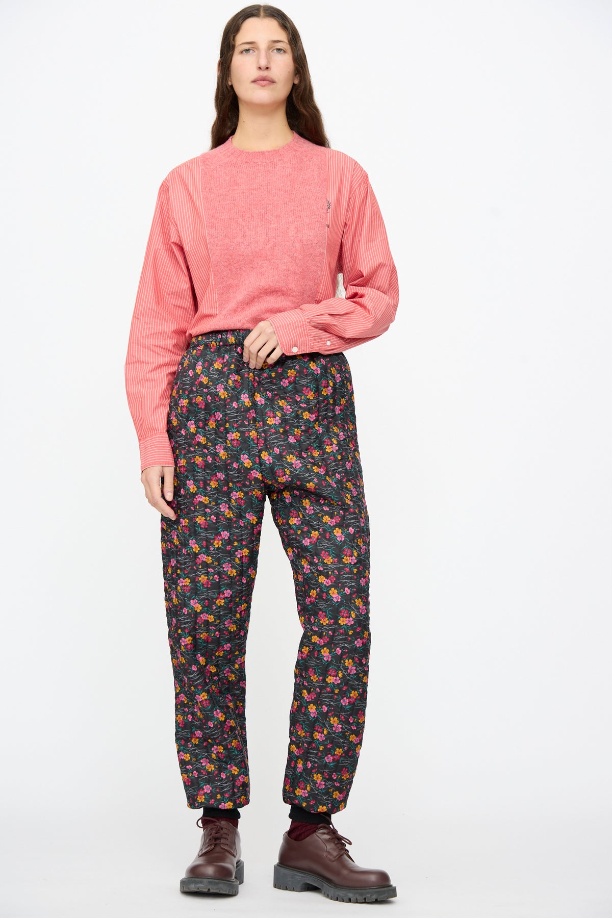 A person wearing a pink striped long-sleeve shirt, Bless No. 08 Monpe Pants in Floral Print IV, and brown lace-up shoes.