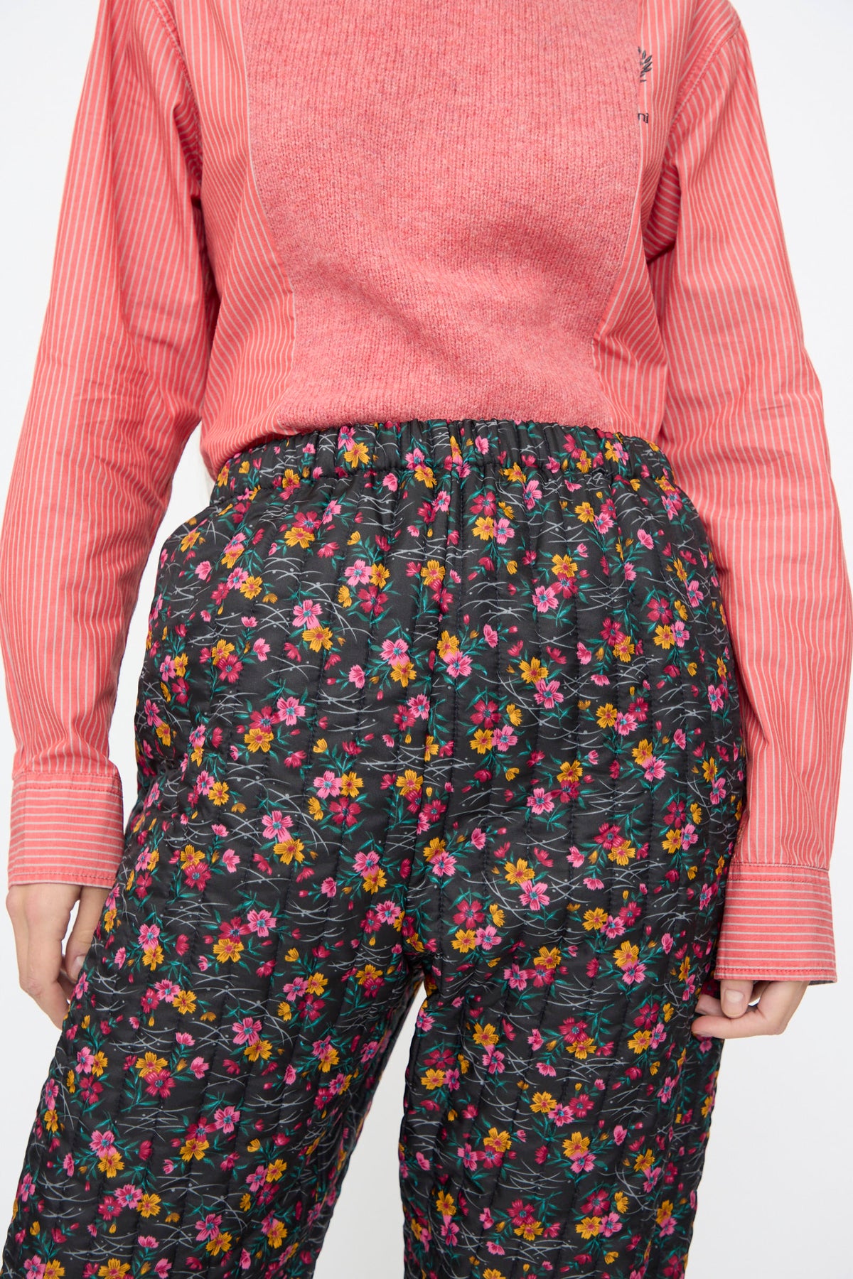 A person wearing a pink striped shirt and Bless No. 08 Monpe Pants in Floral Print IV, which are quilted black floral nylon pants.
