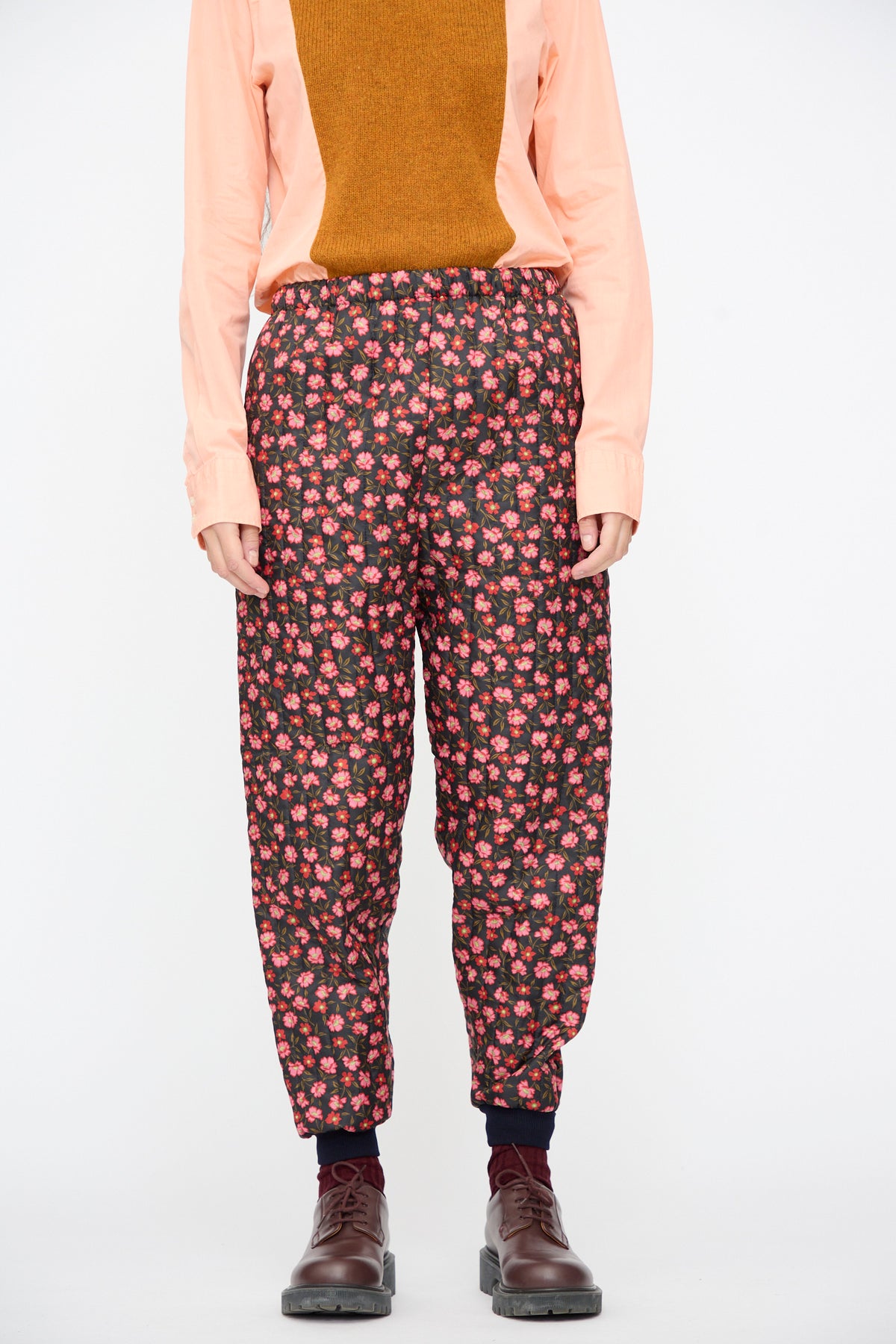 A person wearing Bless No. 08 Monpe Pants in Floral Print V with an elastic waist, a tan and peach long-sleeved shirt, and brown lug-sole shoes.