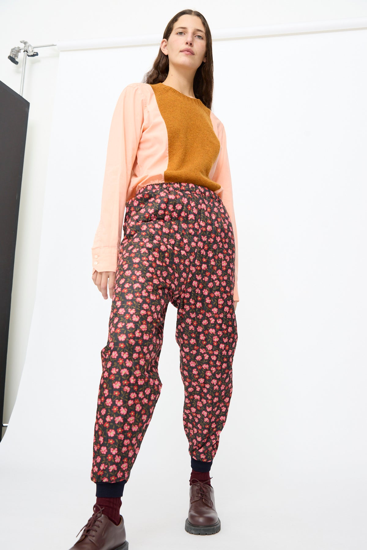 A person wearing a peach and mustard-brown shirt tucked into dark floral Bless No. 08 Monpe Pants in Floral Print V.