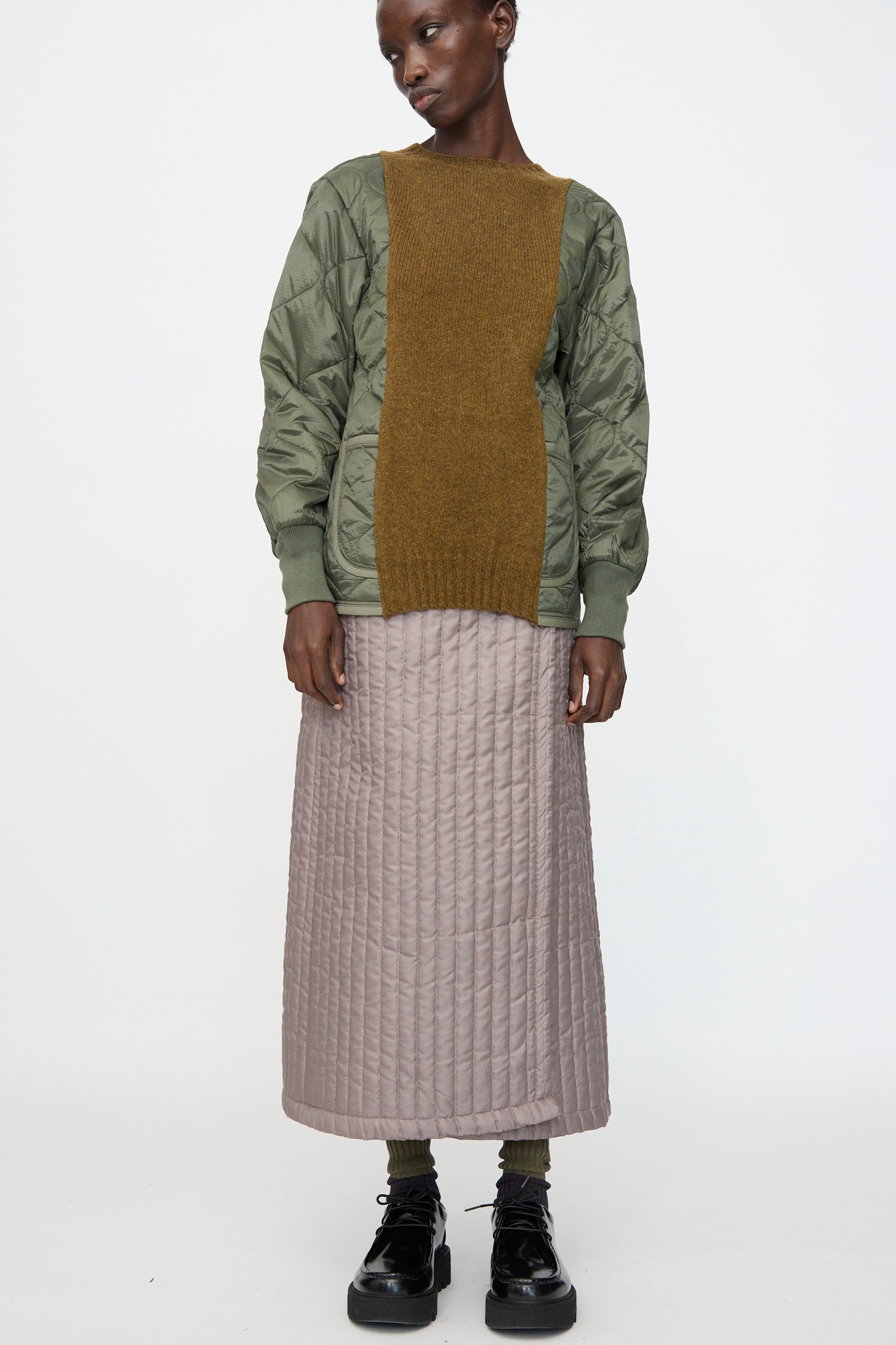 A person wears Bless's No. 08 Monpe Skirt in Beige, paired with a green quilted jacket featuring an olive brown knit panel, dark socks, and black platform shoes against a plain white background.
