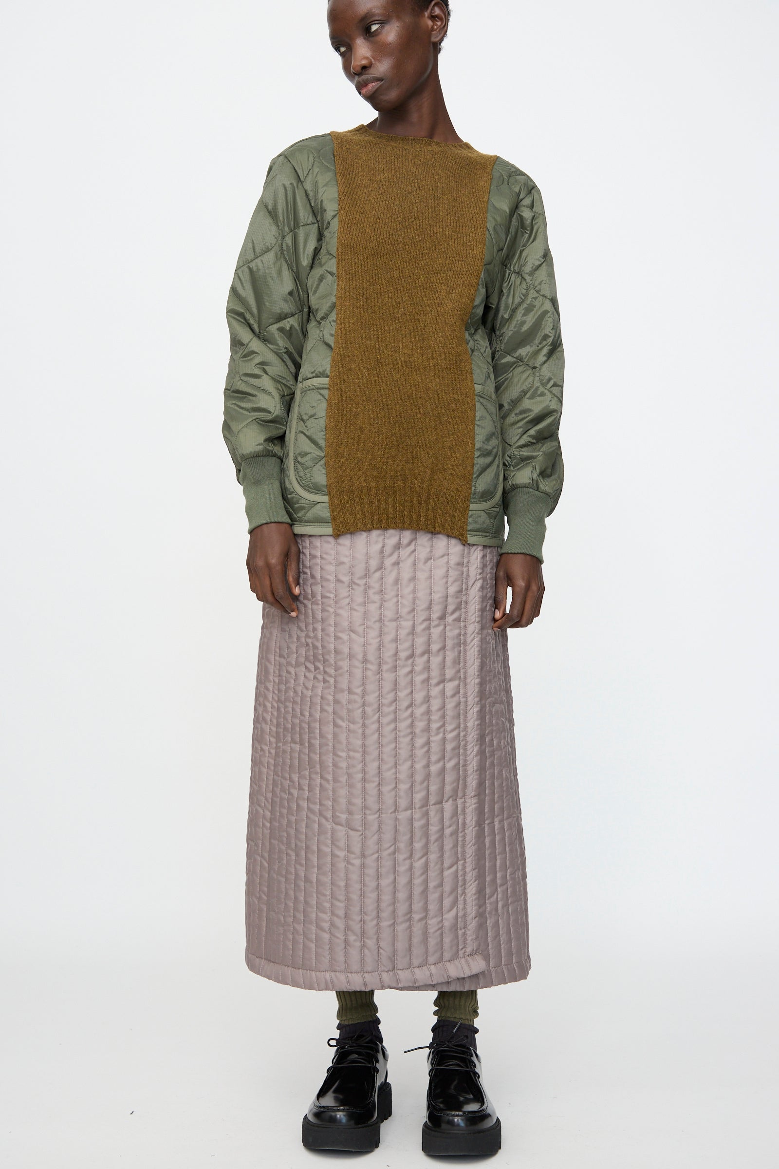 A person wears Bless's No. 08 Monpe Skirt in Beige, paired with a green quilted jacket featuring an olive brown knit panel, dark socks, and black platform shoes against a plain white background.