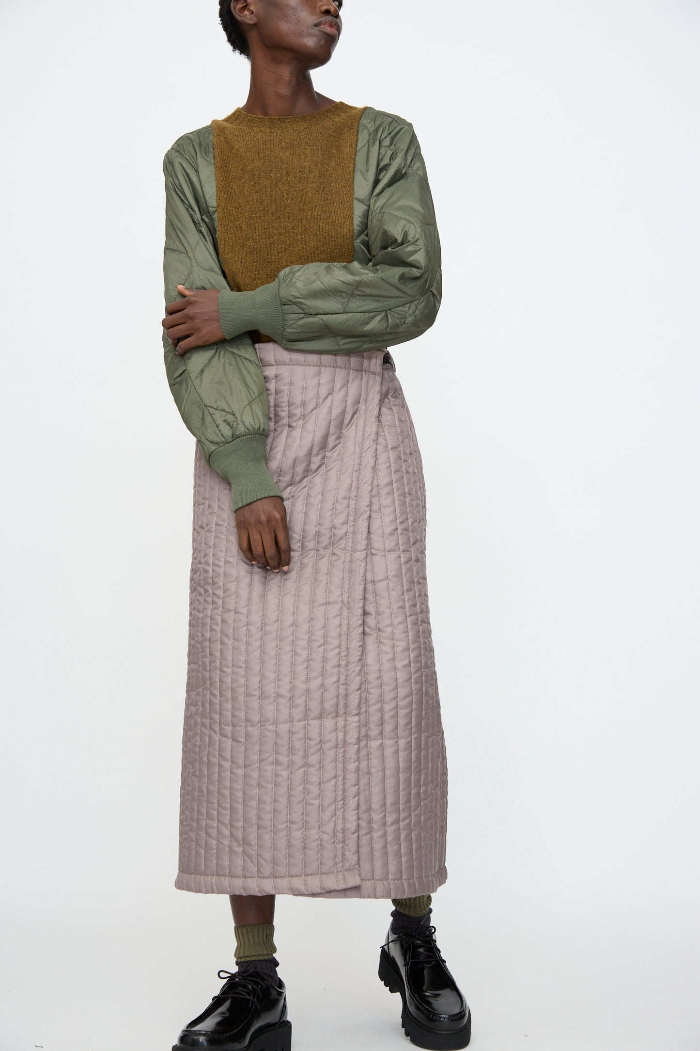 A person stands against a white background wearing a textured green jacket, brown top, Bless No. 08 Monpe Skirt in Beige, and black shoes.