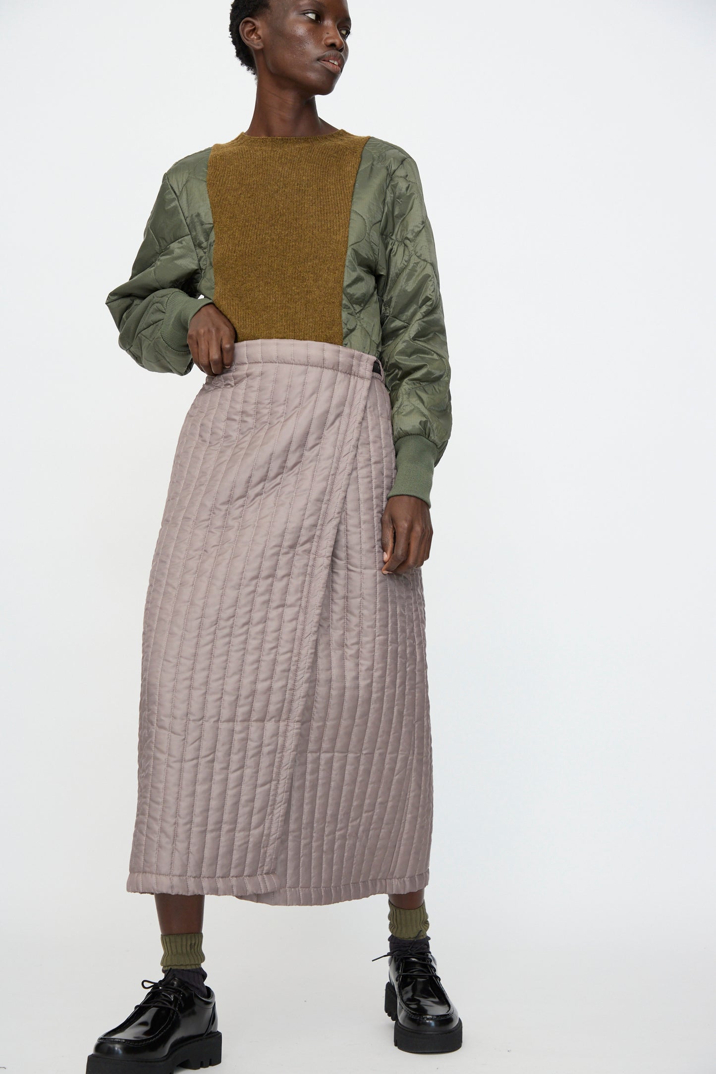Model wears the Bless No. 08 Monpe Skirt in Beige, styled with a textured green and brown top, green socks, and black platform shoes, standing against a plain background.