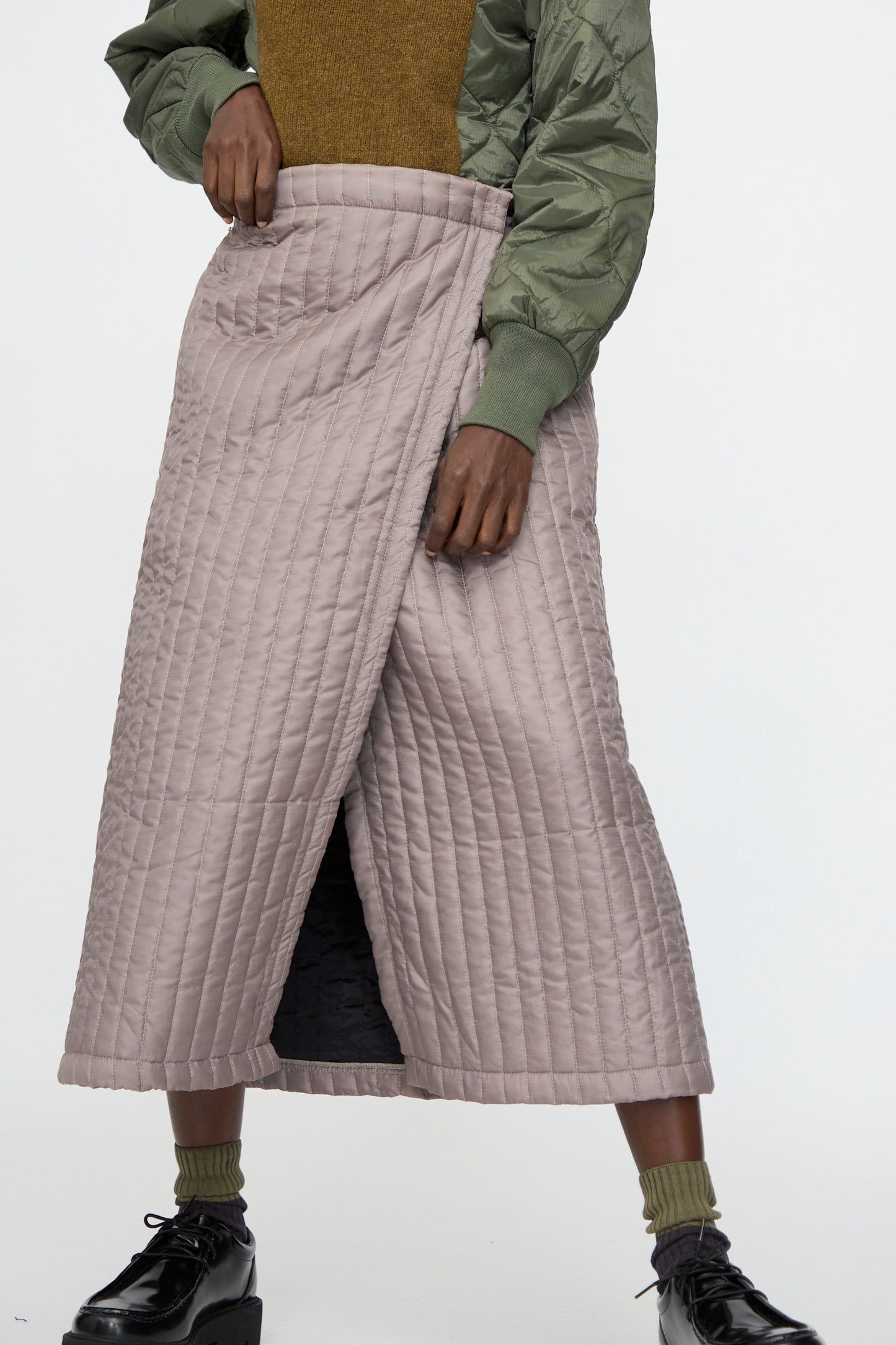 Person wearing the Bless No. 08 Monpe Skirt in Beige (made in Germany), an olive green bomber jacket, mustard top, green socks, and black platform shoes, standing against a plain background.