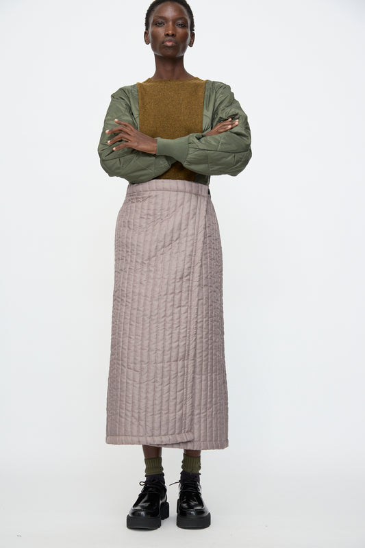 A person stands facing forward wearing a green quilted jacket, brown sweater, Bless No. 08 Monpe Skirt in Beige, green socks, and black platform shoes against a plain white background.