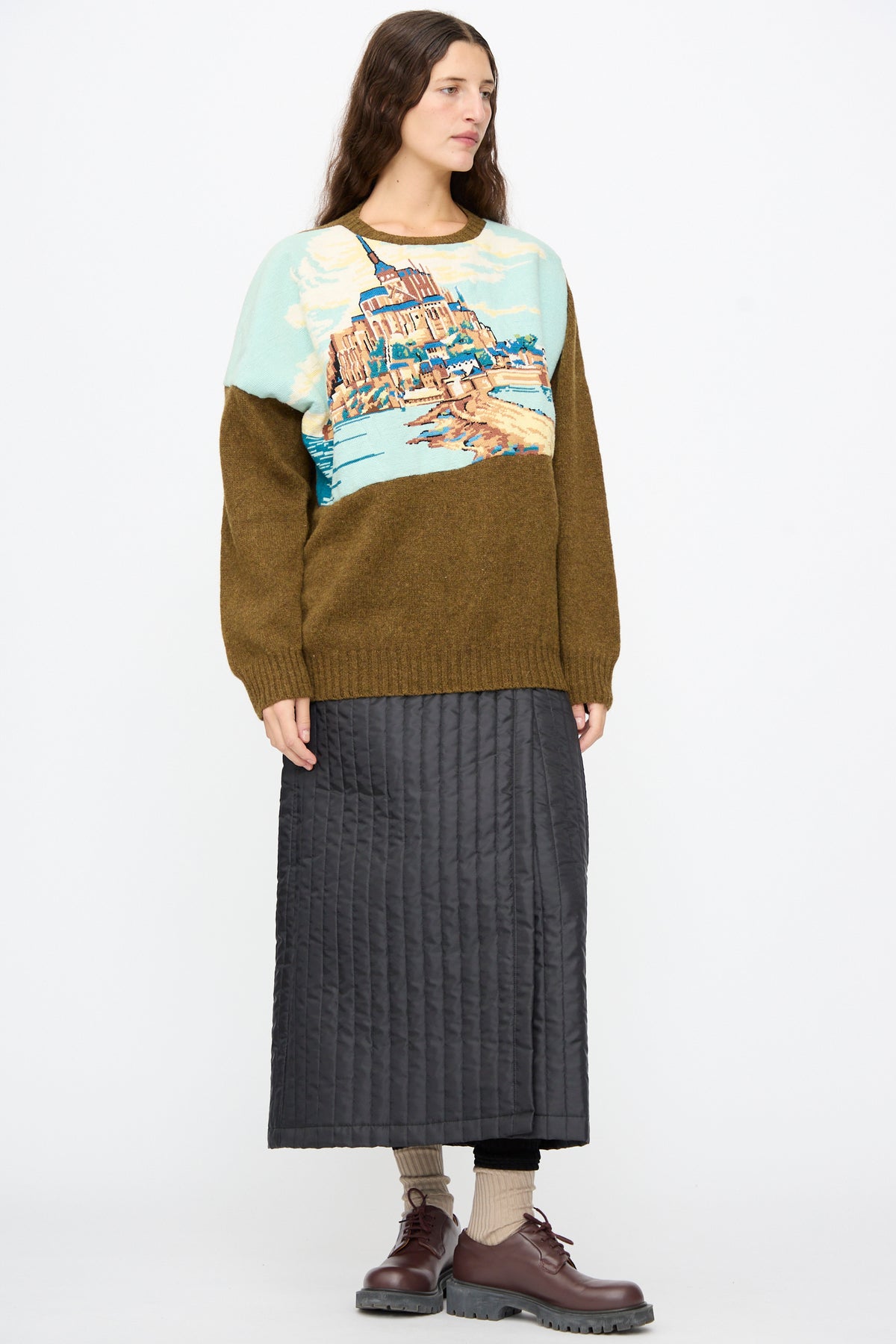 A person wears a long-sleeved sweater featuring a landscape illustration, a black quilted nylon Bless No. 08 Monpe Skirt, and brown leather shoes.