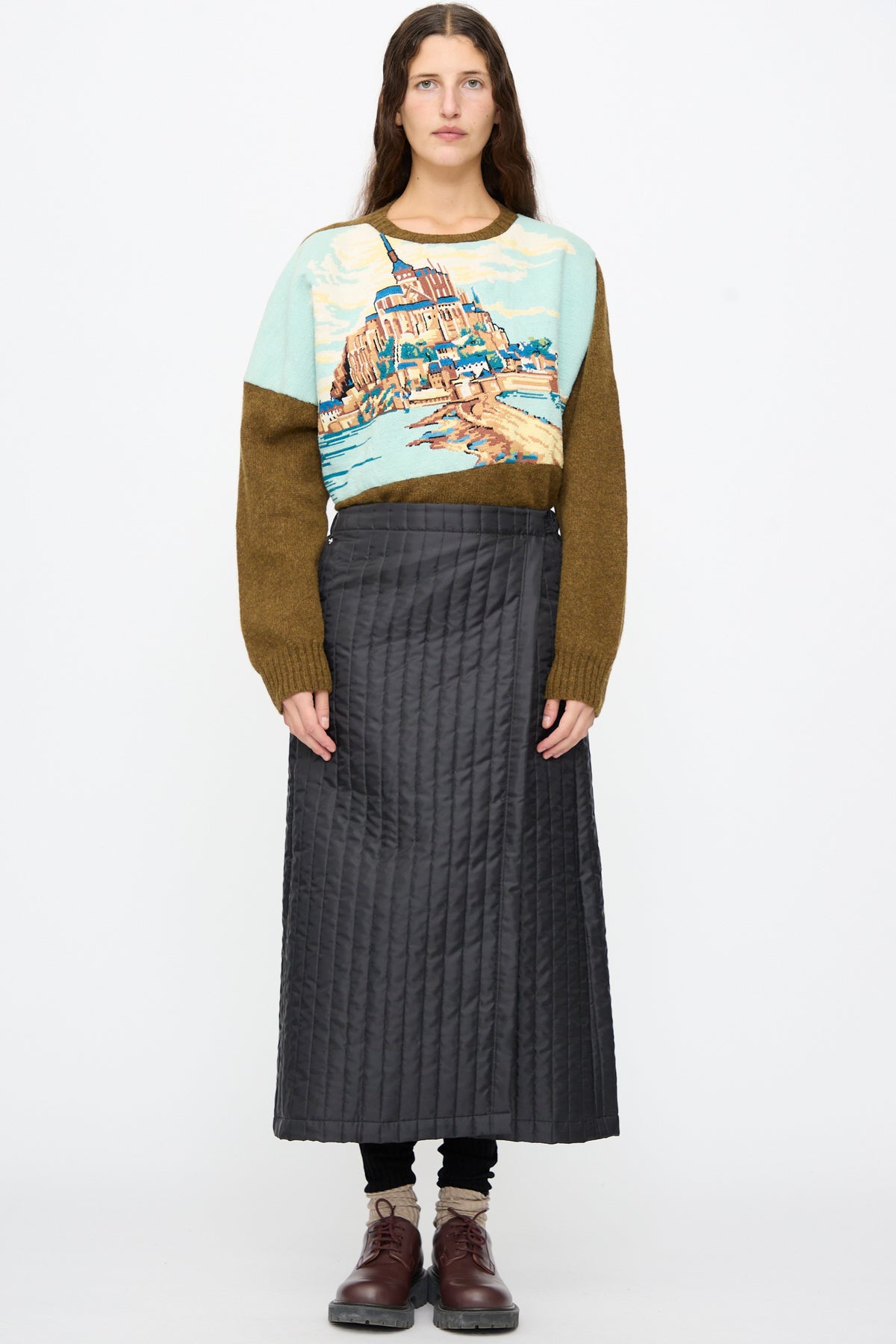 A person wears a color-blocked landscape sweater, a black Bless No. 08 Monpe Skirt in a quilted midi wrap-around style, and brown leather shoes.