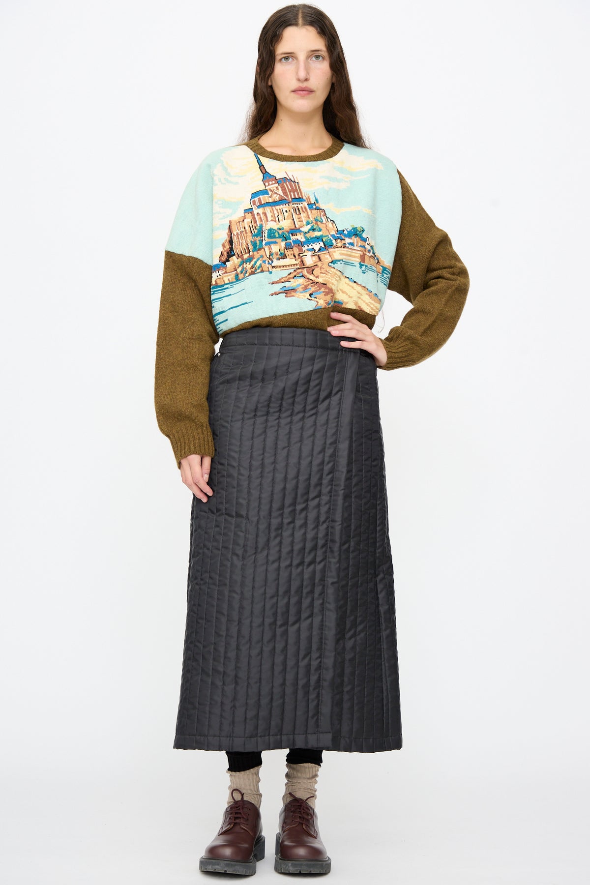 A person wearing a color-blocked sweater featuring an image of Mont Saint-Michel, paired with a black Bless No. 08 Monpe Skirt and dark shoes.