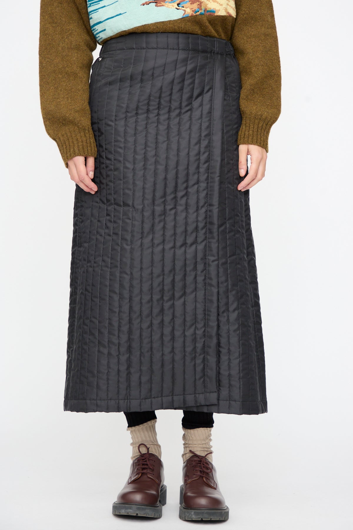 A person wearing a black Bless No. 08 Monpe Skirt in a quilted nylon wrap style, brown sweater, beige socks, and brown shoes.