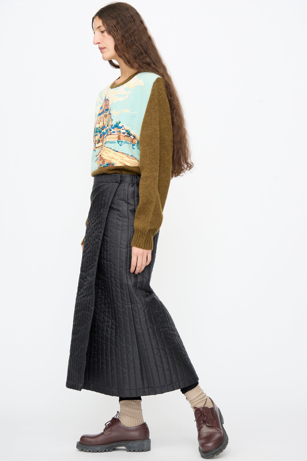 A person wears a black Bless No. 08 Monpe Skirt, a landscape-print sweater, beige socks, and dark brown lace-up shoes.