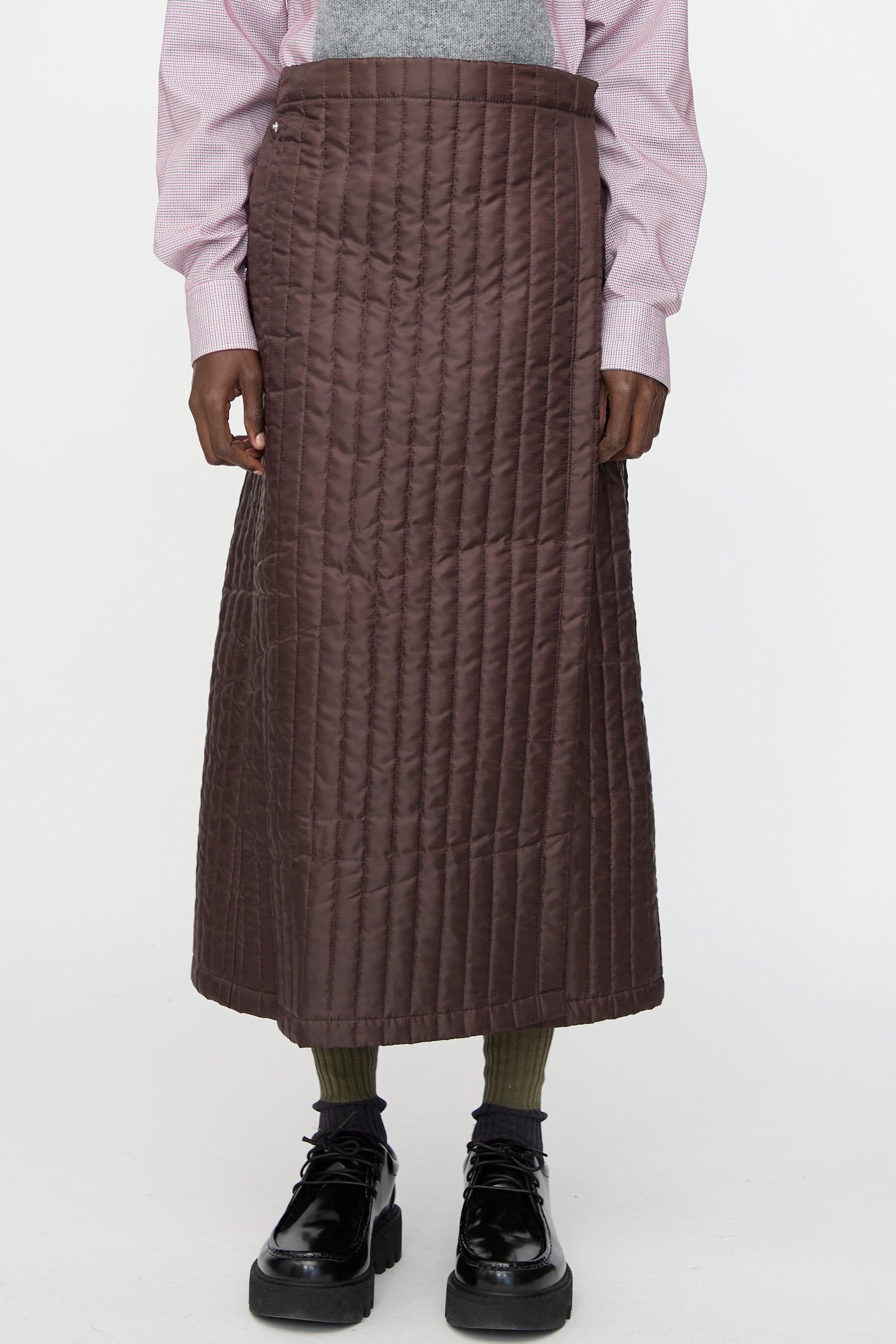 A person wears the Bless No. 08 Monpe Skirt in Brown, paired with a pink long-sleeve shirt, olive socks, and black shiny shoes, standing against a plain white background.