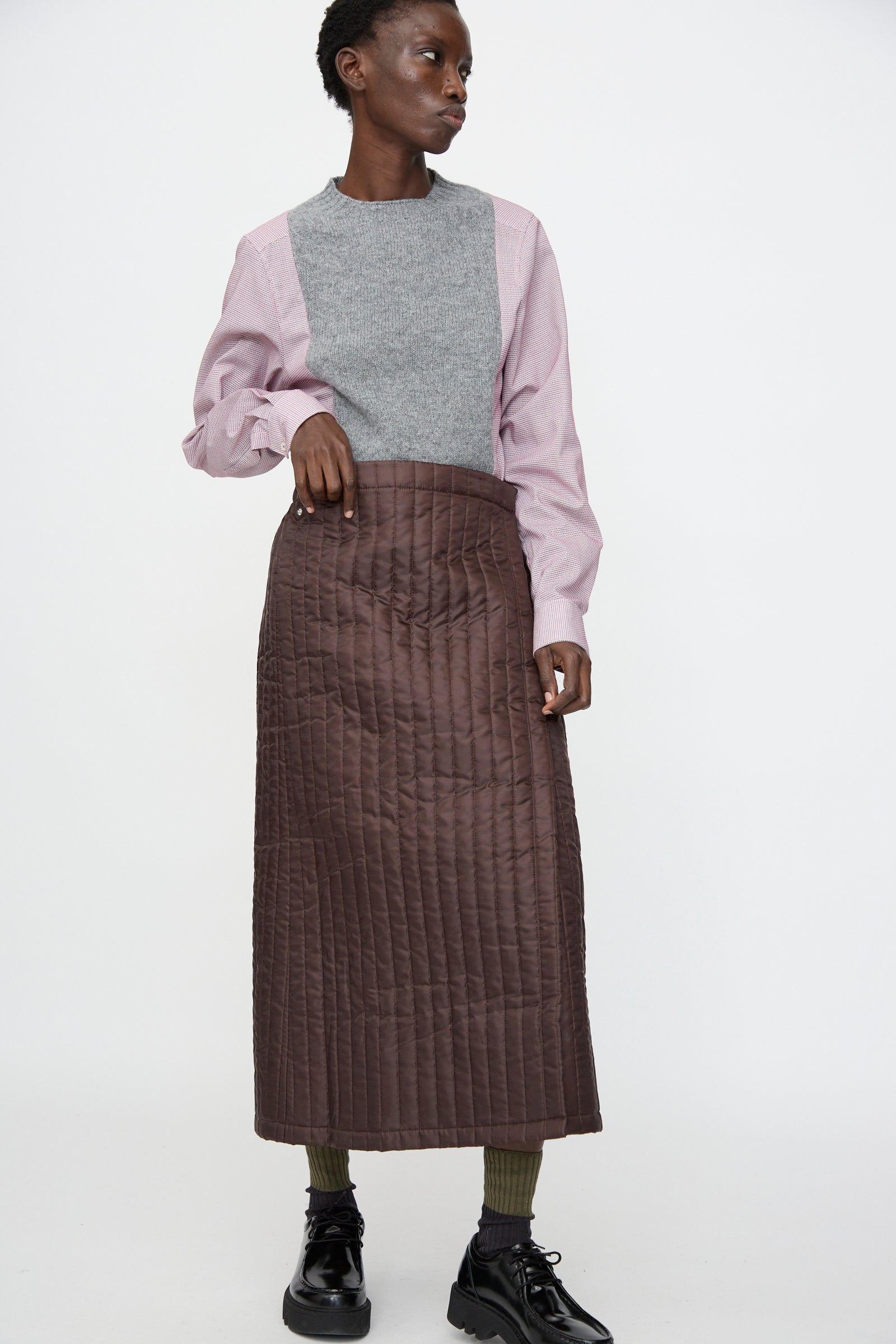 A person wears the Bless No. 08 Monpe Skirt in Brown with a gray and pink long-sleeve top, olive green socks, and black shoes, standing against a plain white background.