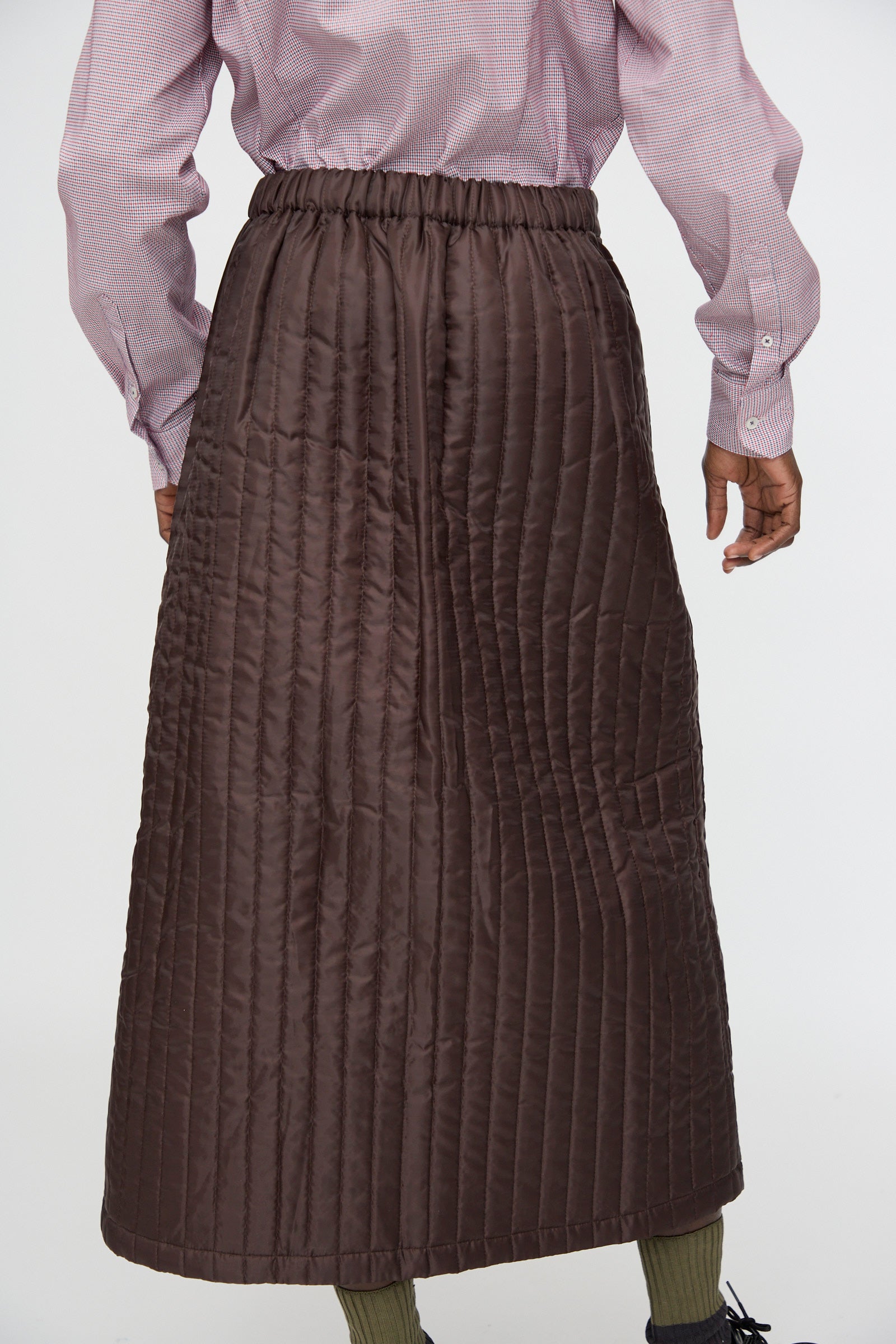 A person is seen from behind wearing the Bless No. 08 Monpe Skirt in Brown with vertical quilting, a light pink long-sleeve shirt, and green ribbed socks.