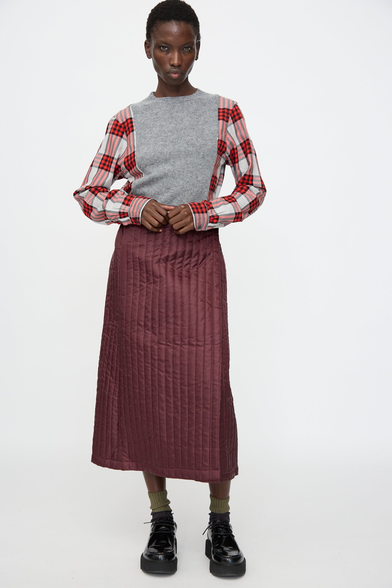 Person standing against a plain background, wearing a gray sweater over a red plaid shirt, the No. 08 Monpe Skirt in Burgundy by Bless (made in Germany), olive socks, and black shoes.