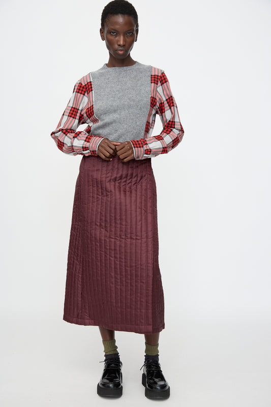 Person standing against a plain background, wearing a gray sweater over a red plaid shirt, the No. 08 Monpe Skirt in Burgundy by Bless (made in Germany), olive socks, and black shoes.