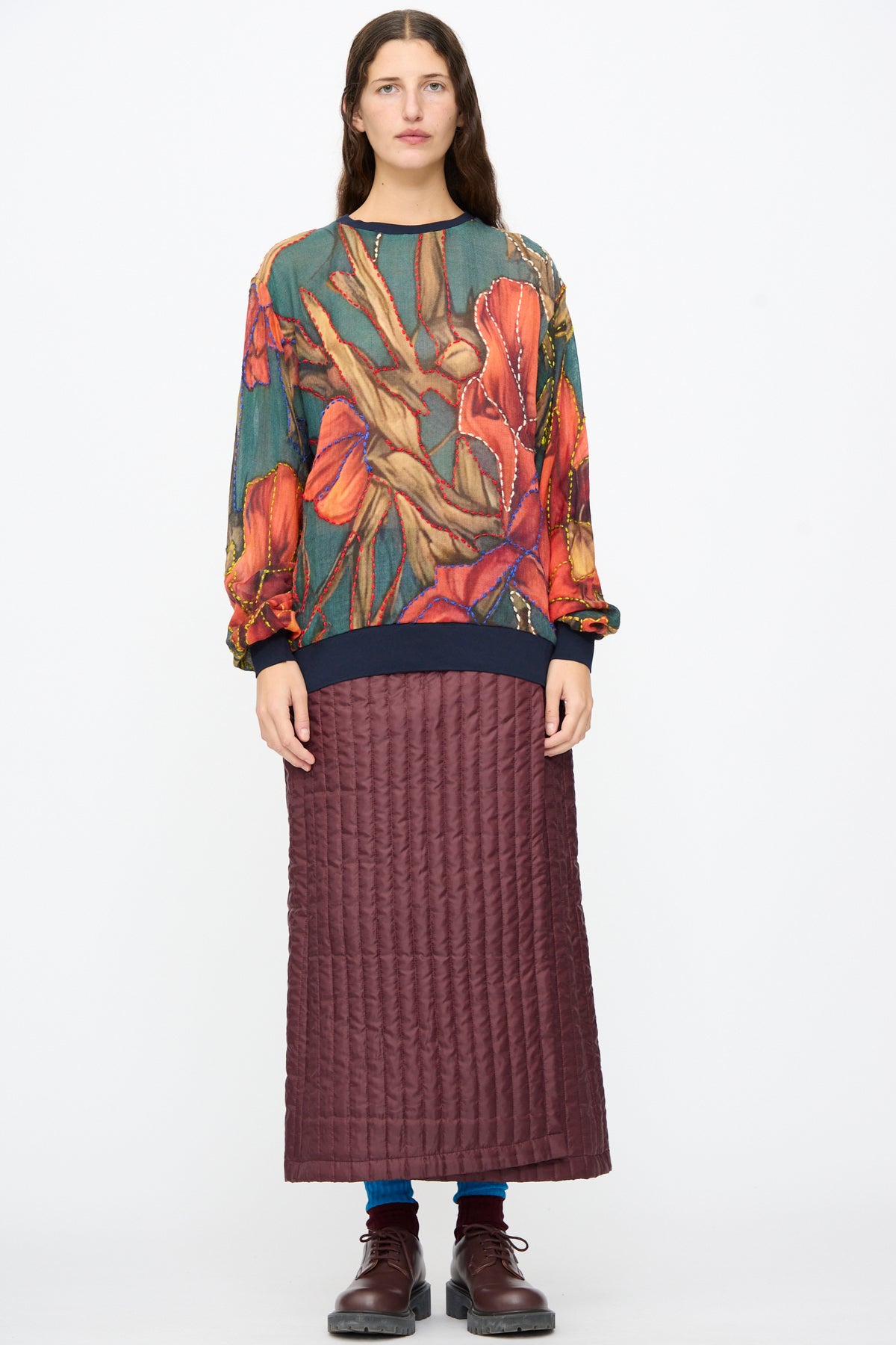 A person wears a colorful botanical print top and a burgundy Bless No. 08 Monpe Skirt in a quilted nylon midi wrap-around style, paired with dark boots and blue socks.