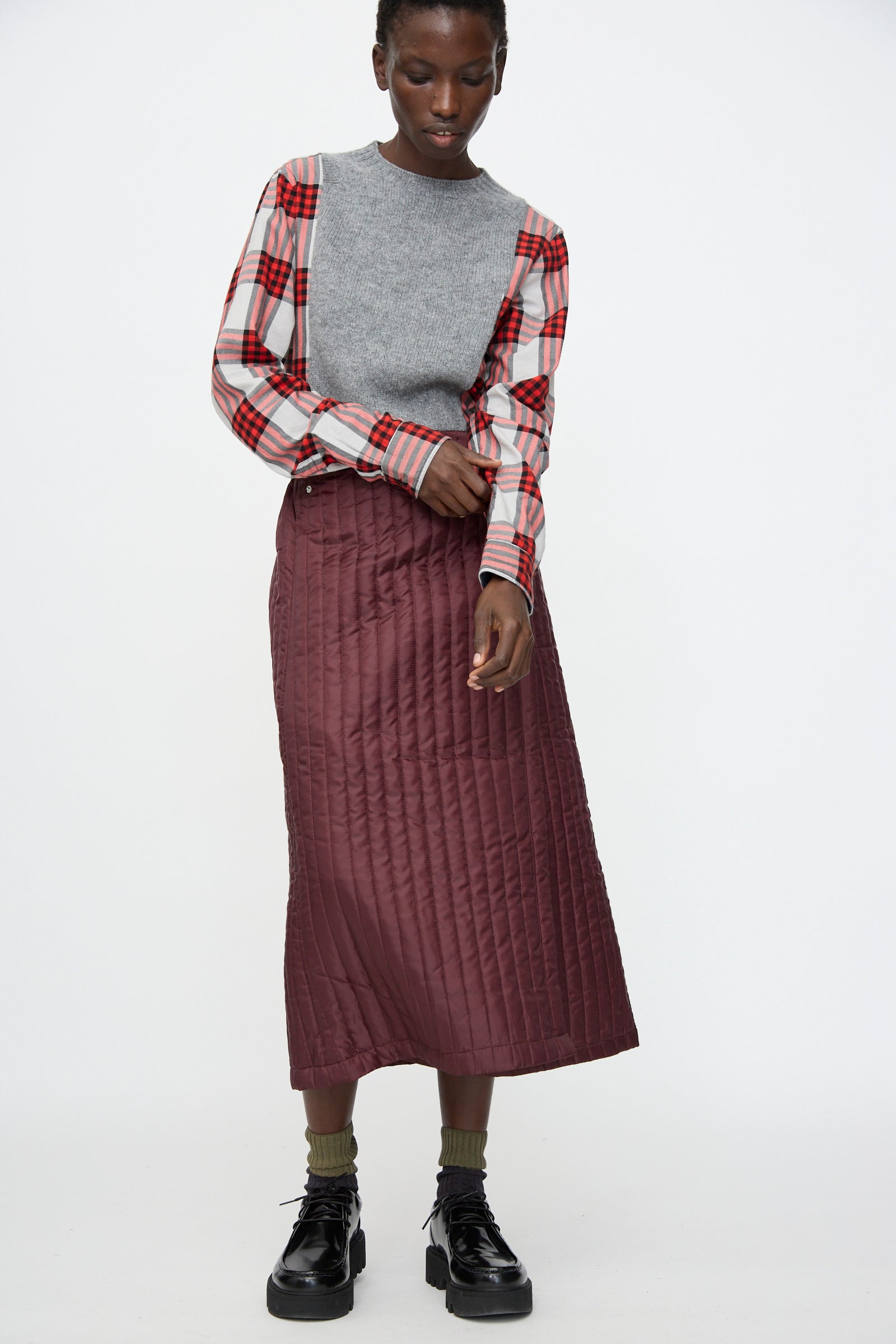 A person in a gray sweater over a red plaid shirt, the Bless No. 08 Monpe Skirt in Burgundy, olive socks, and black platform shoes stands against a plain white background.