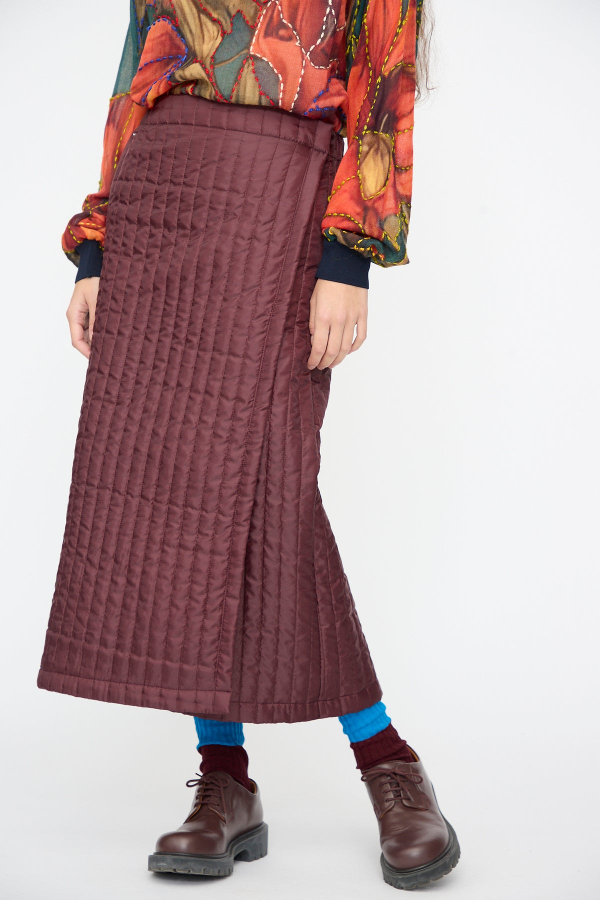 A person wears a Bless No. 08 Monpe Skirt in Burgundy over blue leggings and dark brown lace-up shoes.