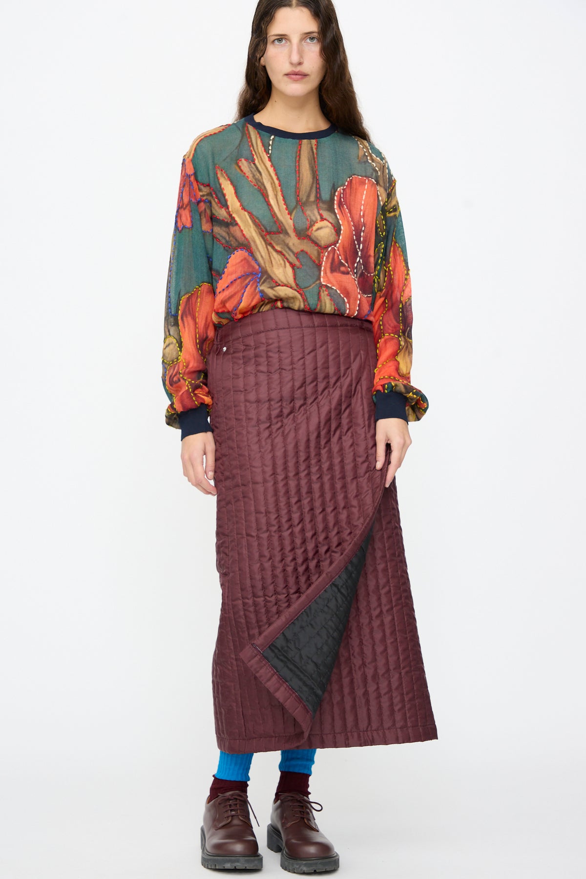 A person wears a burgundy Bless No. 08 Monpe Skirt in a quilted midi wrap-around style over blue leggings, paired with a patterned long-sleeved top and brown lace-up shoes.