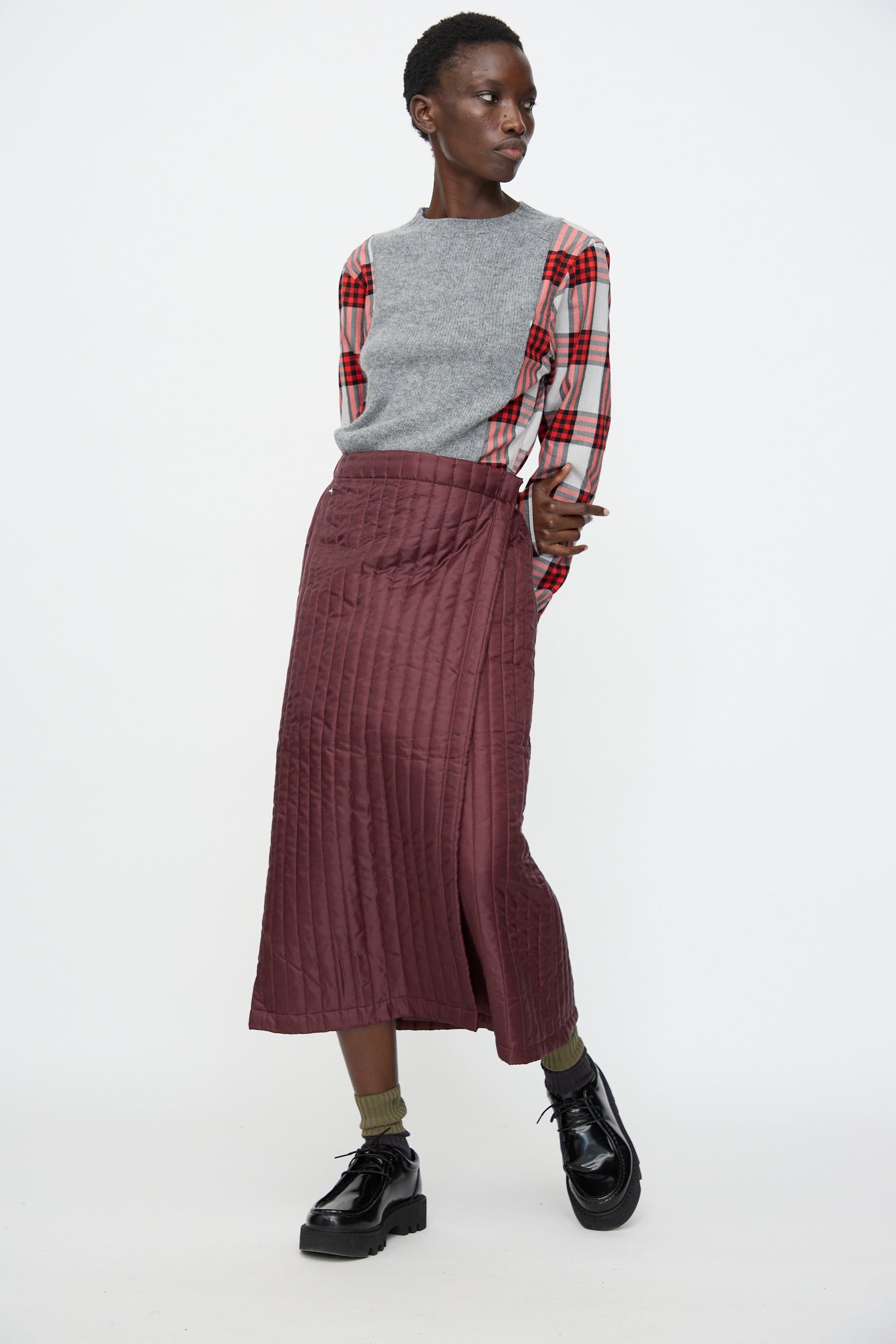 A person stands against a plain white background, wearing a grey vest over a red plaid shirt, the Bless No. 08 Monpe Skirt in Burgundy, olive socks, and black platform shoes.