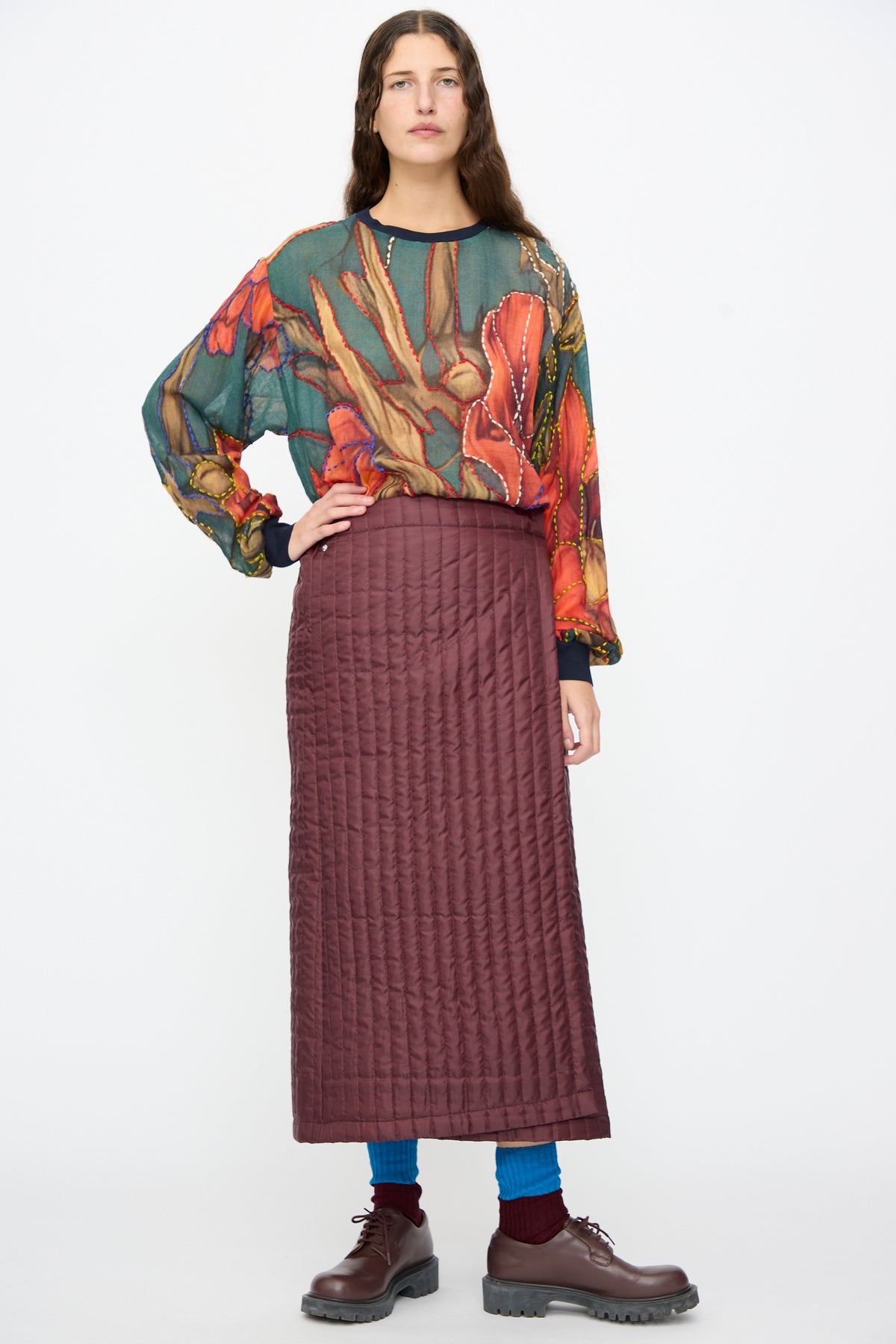 A person wearing a burgundy Bless No. 08 Monpe Skirt, which is a long, quilted nylon midi wrap-around skirt, styled with a patterned long-sleeve top, bright blue socks, and dark brown loafers.