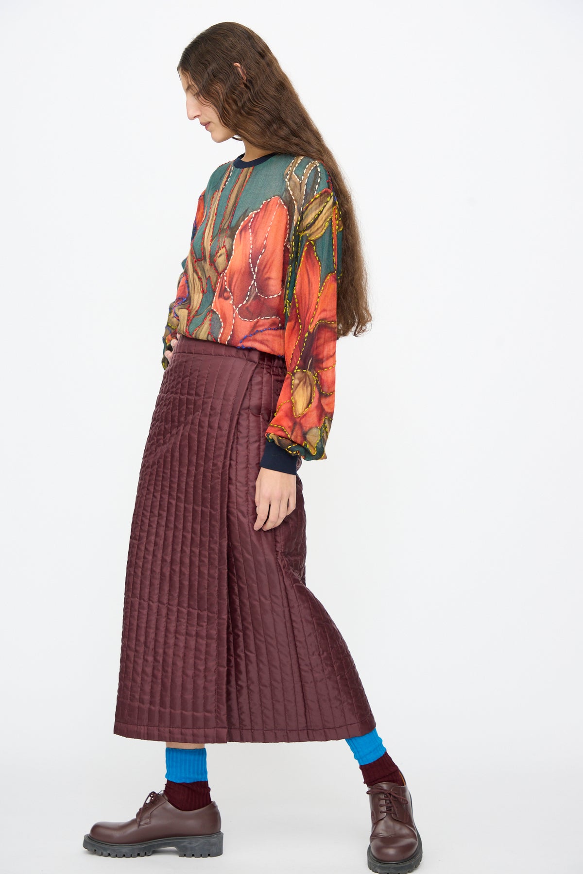 A person wearing a Bless No. 08 Monpe Skirt in Burgundy, a quilted midi wrap-around skirt, styled with a colorful abstract print long-sleeved top, blue socks, and brown lace-up shoes.