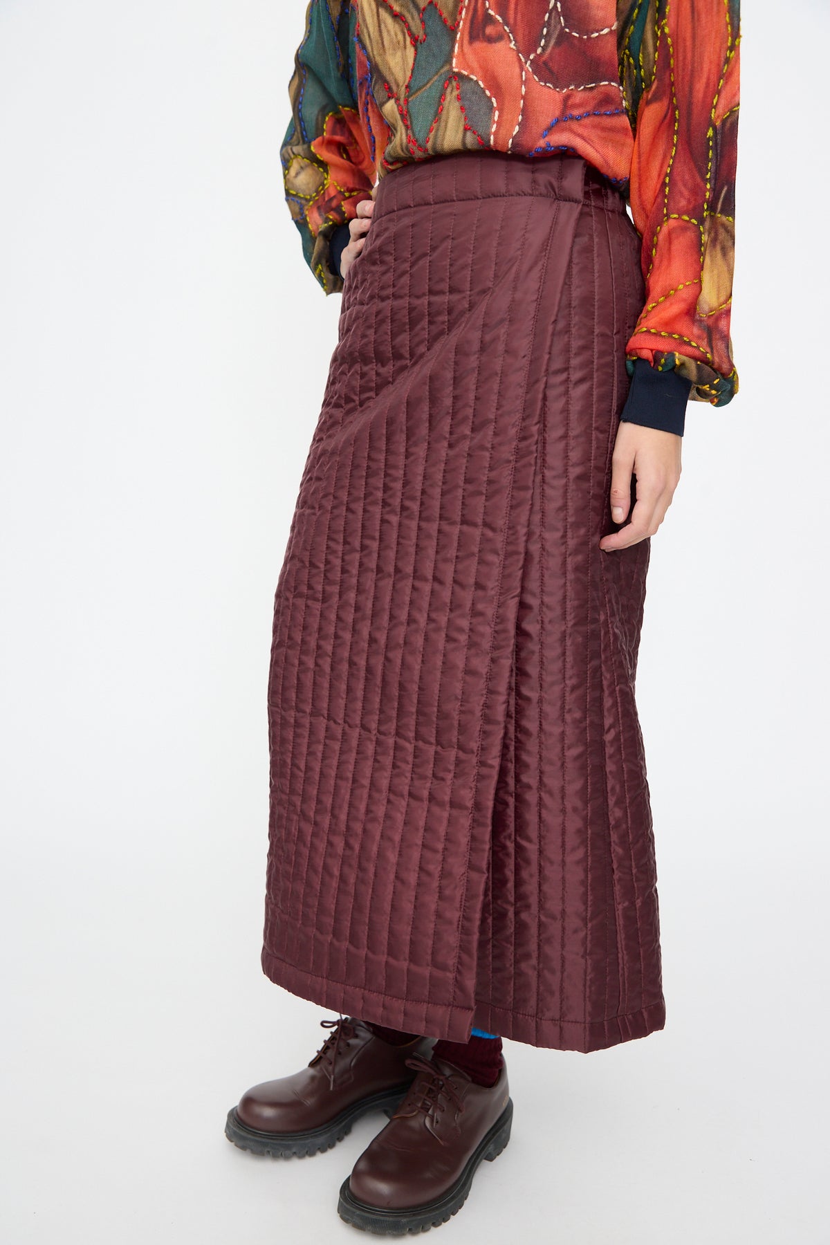 Bless No. 08 Monpe Skirt in burgundy quilted nylon midi wrap style, worn with a colorful patterned long-sleeve top and brown lace-up shoes.