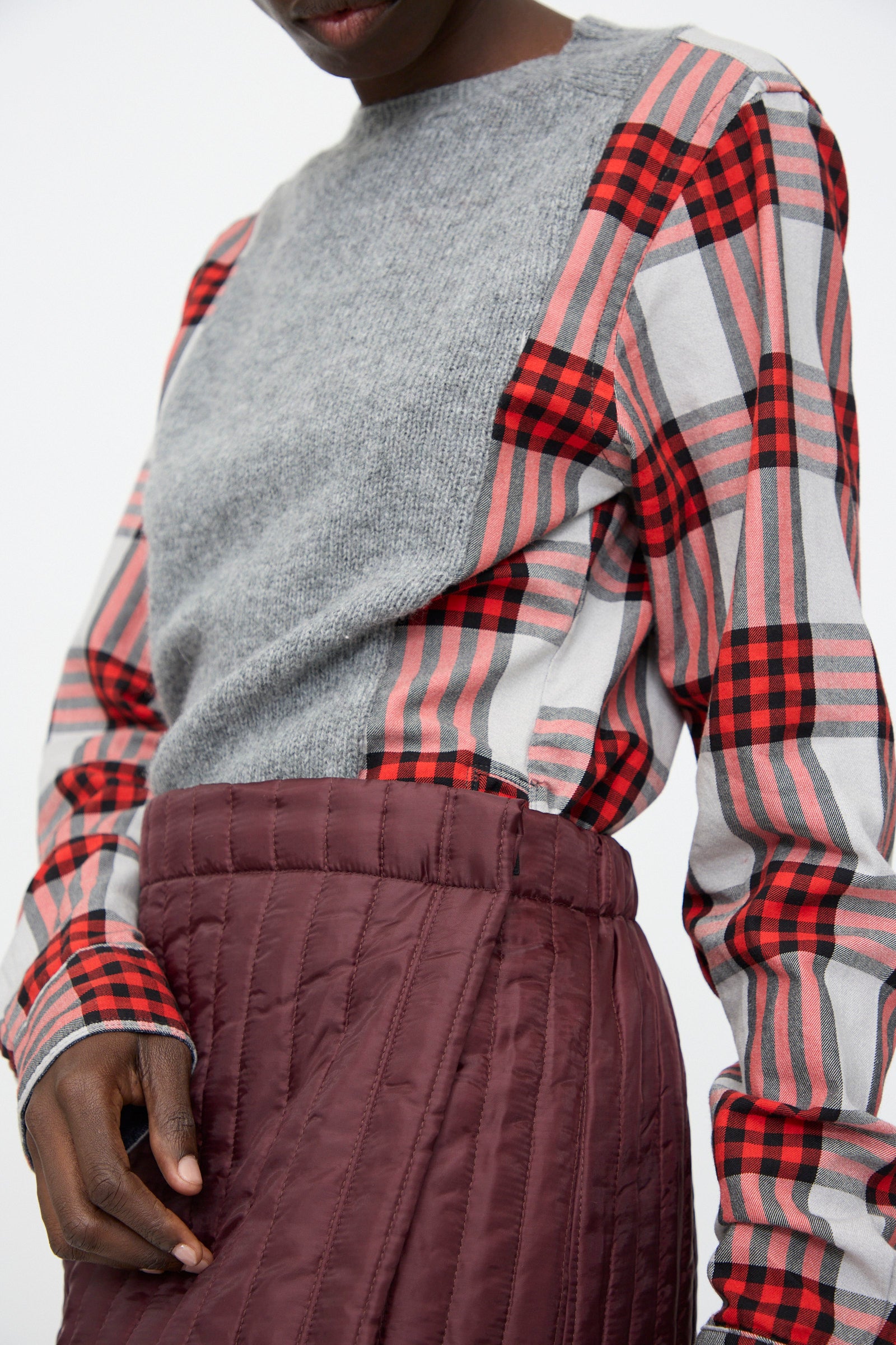A person wearing a gray top with red and black plaid sleeves, paired with the Bless No. 08 Monpe Skirt in Burgundy—a high-waisted, quilted nylon skirt made in Germany.