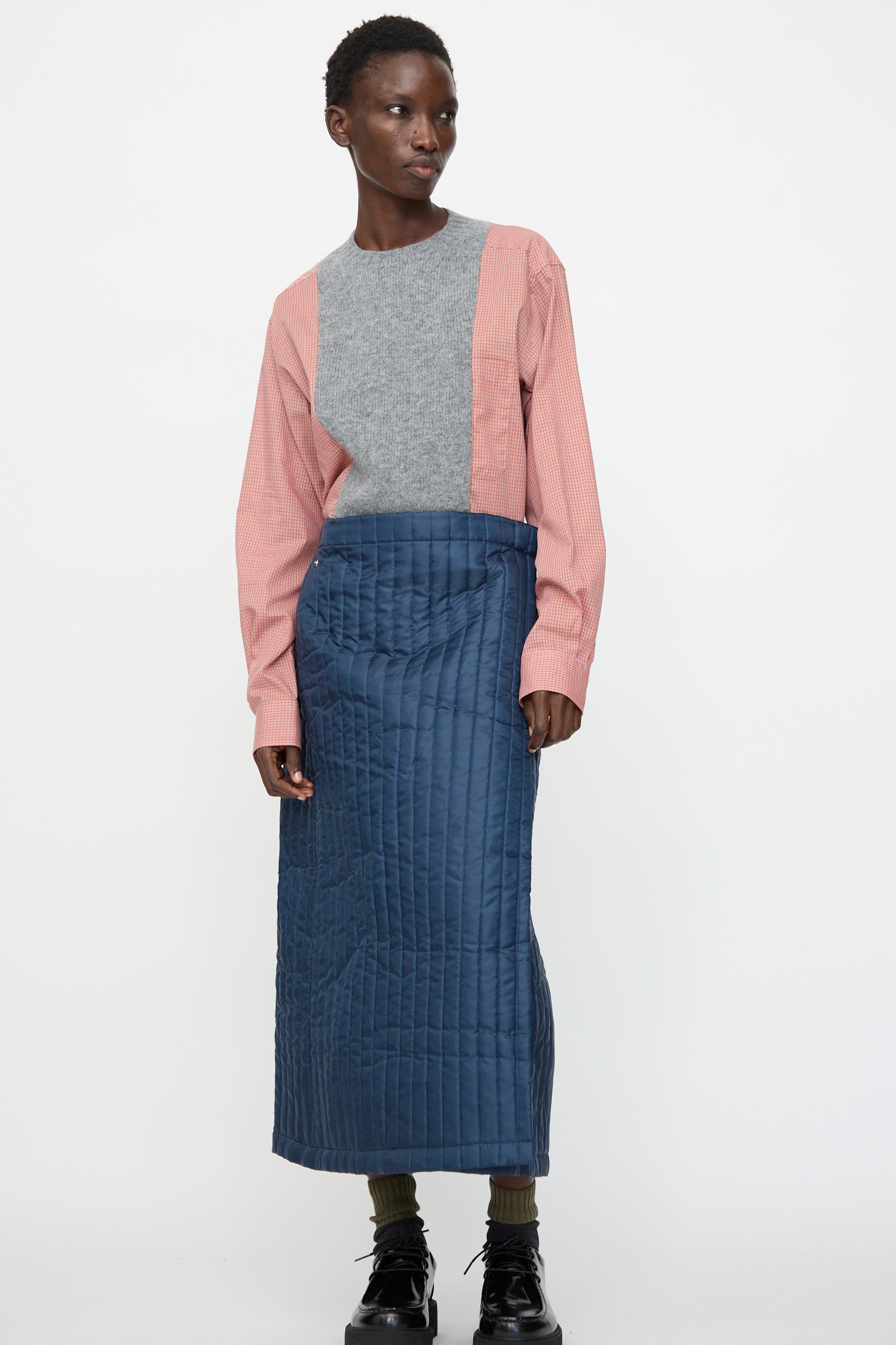 A person stands against a plain white background, wearing a pink and gray long-sleeve top, the Bless No. 08 Monpe Skirt in Navy, green socks, and black shoes.