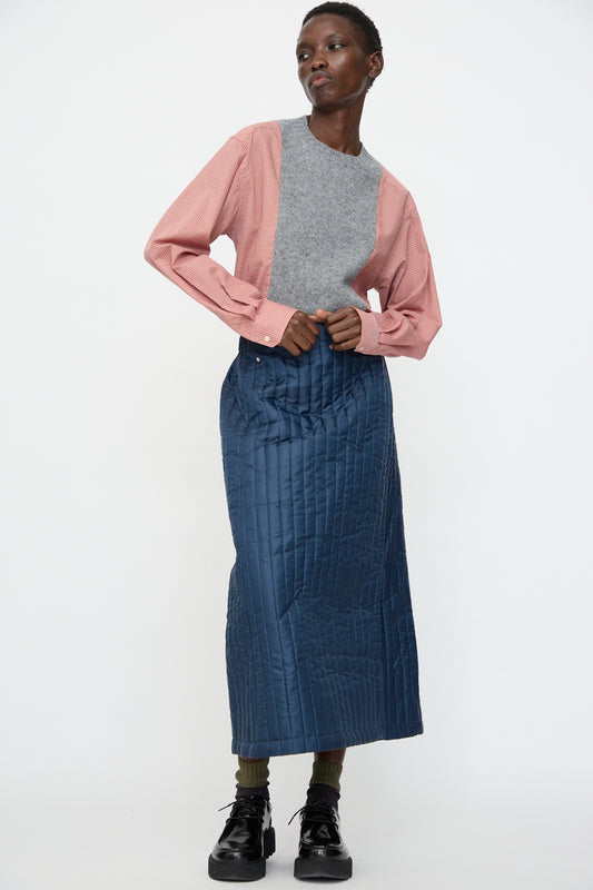 A person stands against a plain white background wearing a gray and pink long-sleeve top, Bless’s No. 08 Monpe Skirt in Navy—a blue quilted nylon skirt with an adjustable snap waist—paired with green socks and black shoes.