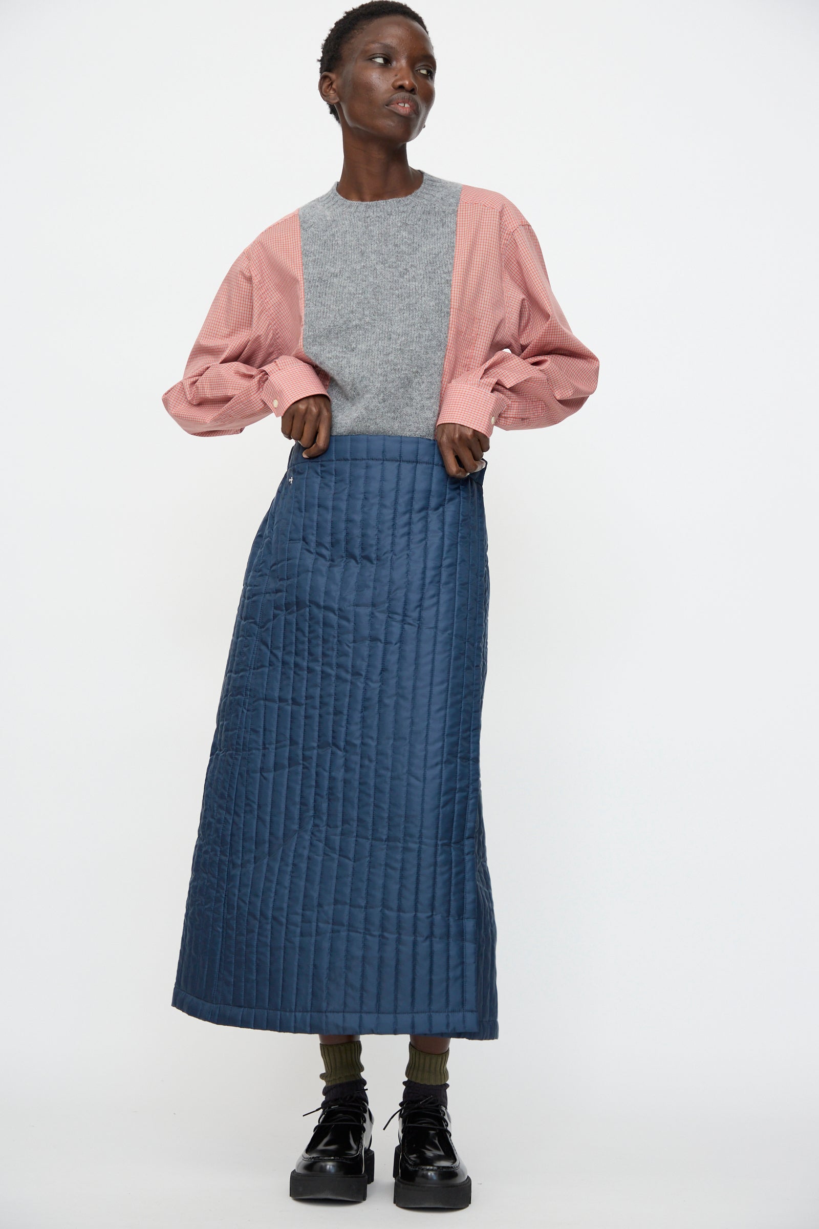 A person wearing a gray and pink long-sleeve top, Bless’s No. 08 Monpe Skirt in Navy with an adjustable snap waist, green socks, and black platform shoes stands against a plain white background.