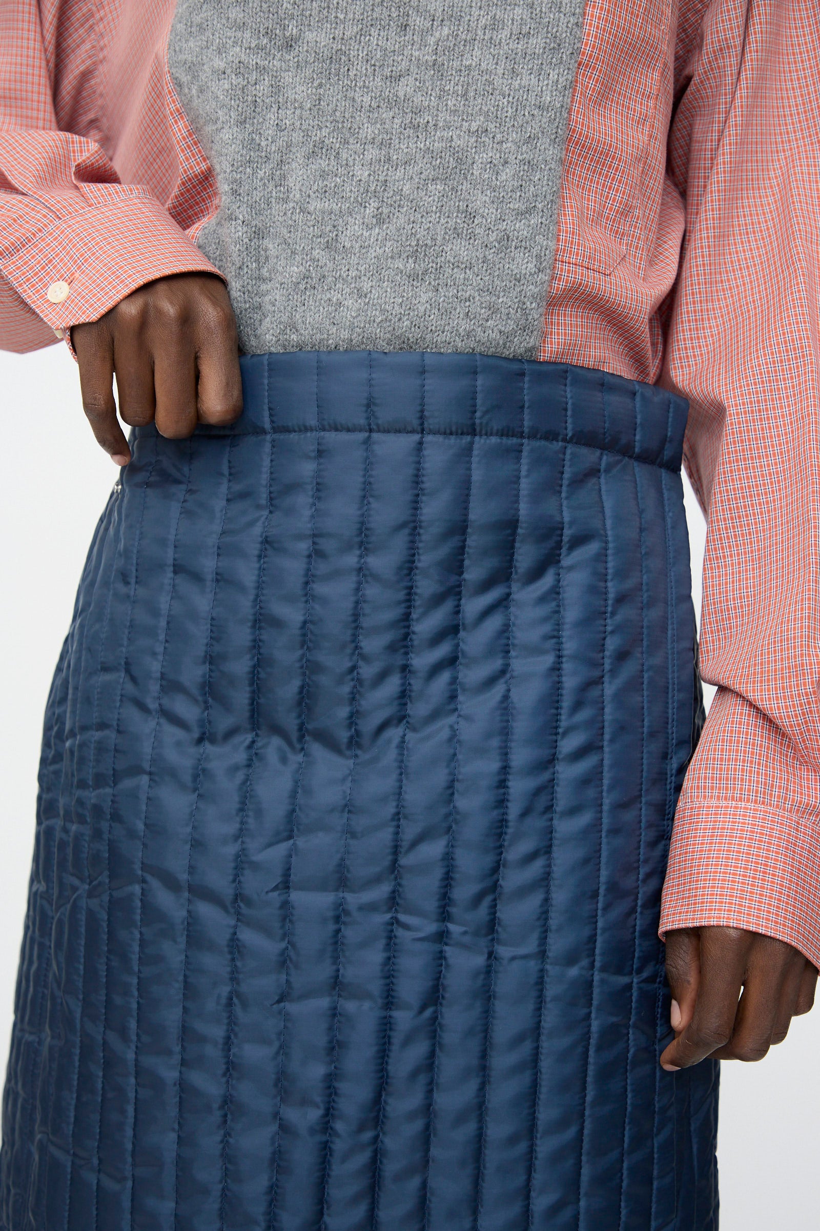 A person wears a grey sweater, a pink checkered shirt, and the Bless No. 08 Monpe Skirt in Navy, with one hand resting on the skirt’s waistband.