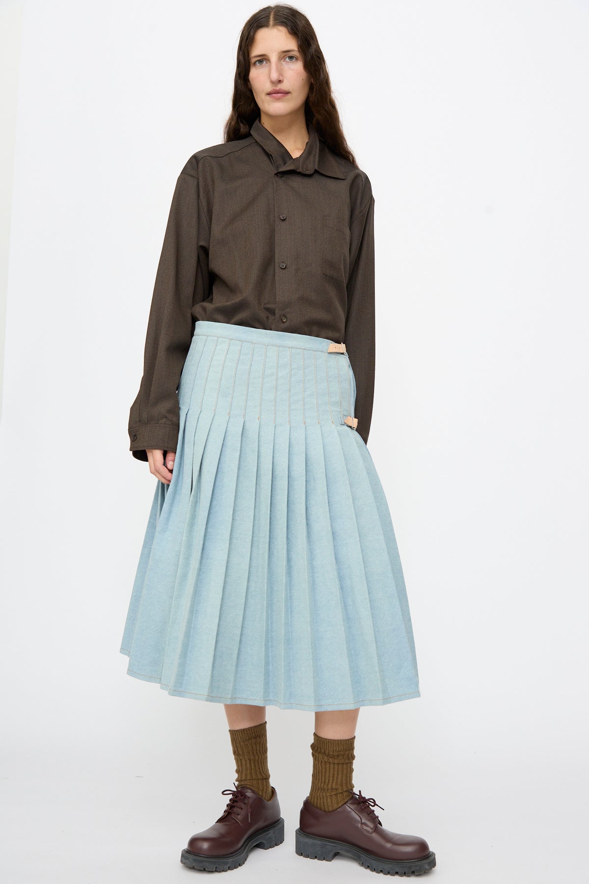 A person wears a dark button-down shirt and a Bless No. 52 Denimkilt in Denim Light Blue featuring side buckle straps, paired with brown socks and dark brown lace-up shoes.