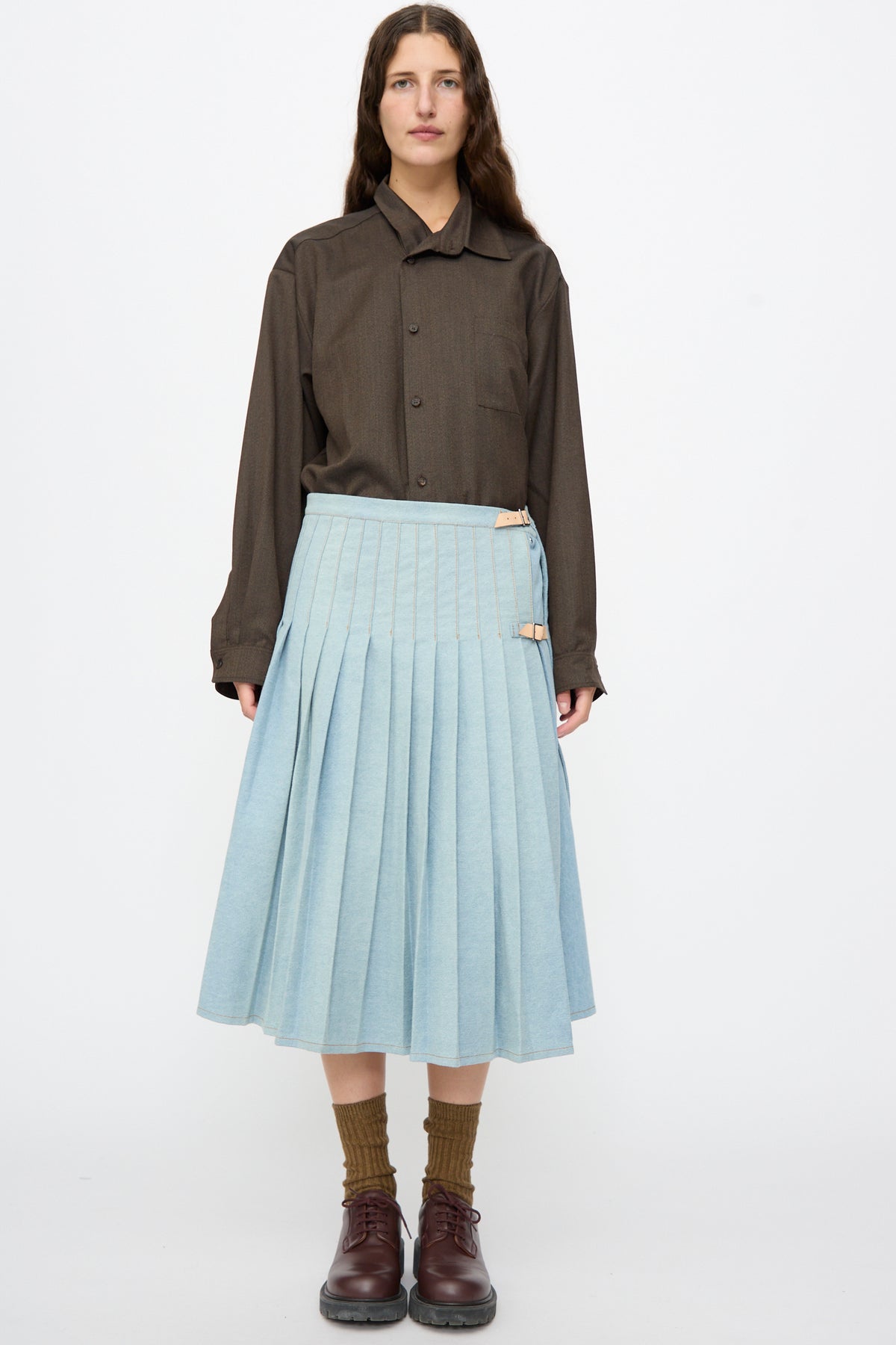 A person wearing a long-sleeved dark brown shirt and a Bless No. 52 Denimkilt in Denim Light Blue, a pleated midi skirt with buckled side straps.