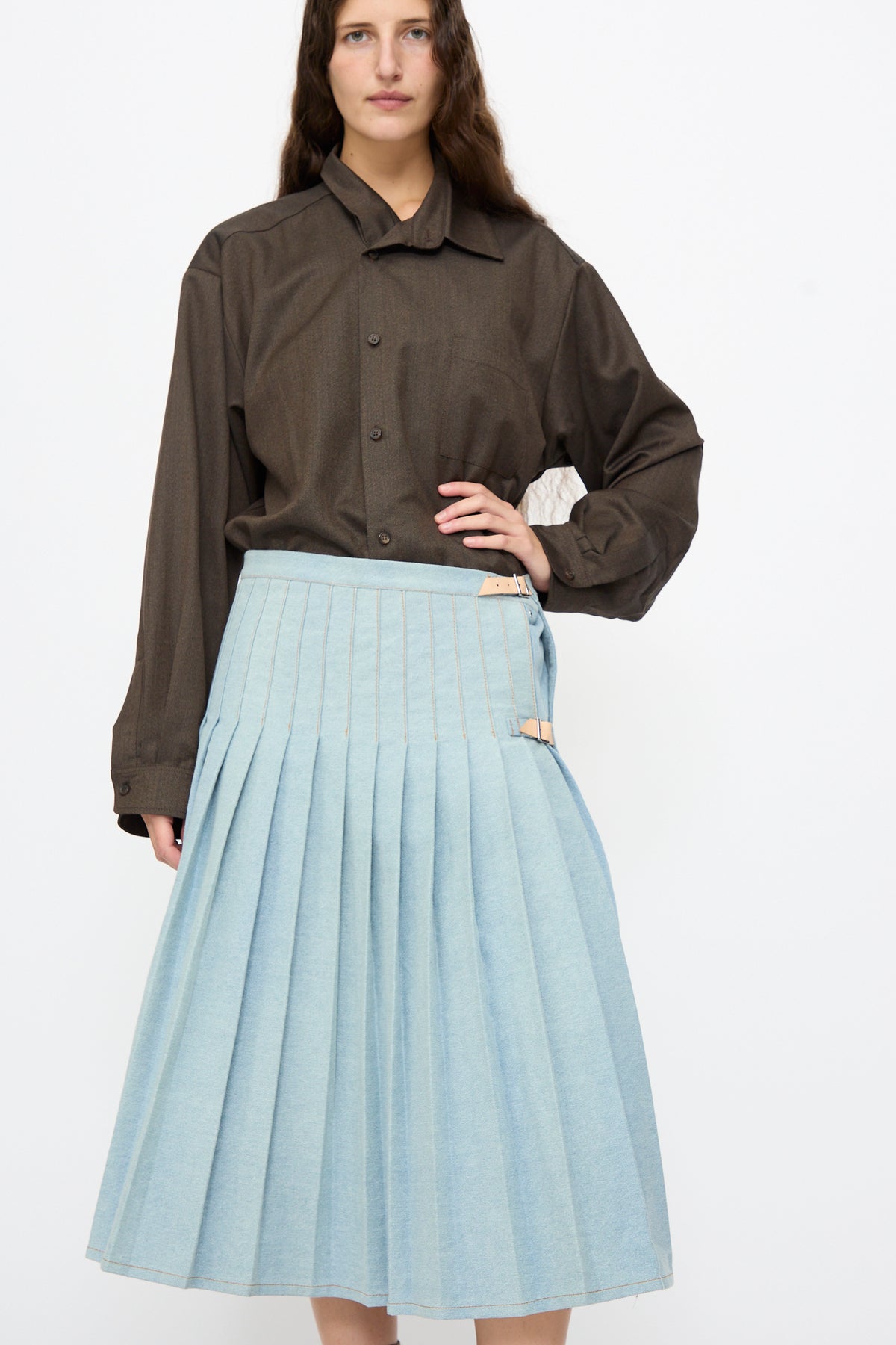 A person wears a long-sleeved dark brown button-down shirt tucked into a Bless No. 52 Denimkilt in Denim Light Blue, a pleated kilt with side buckle adjustments.