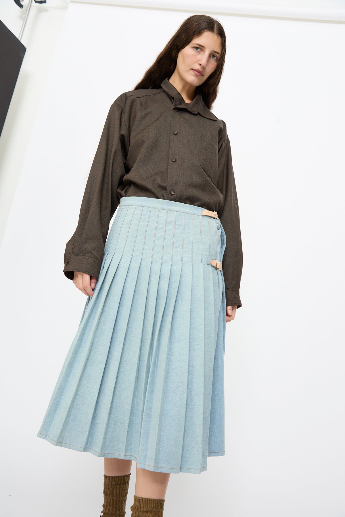 A person wears a light blue pleated Bless No. 52 Denimkilt in Denim Light Blue with buckled side straps and a dark brown long-sleeved shirt.