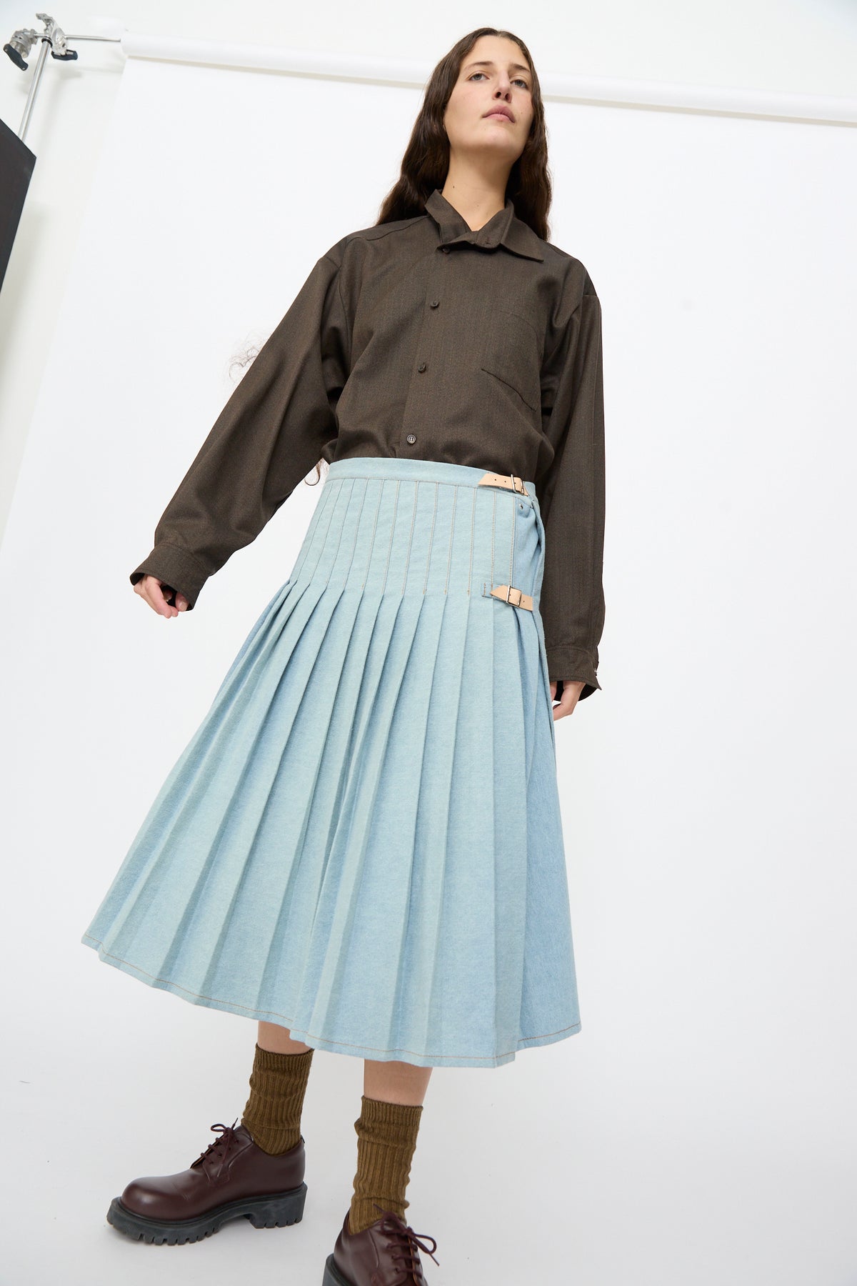 A person wearing a long-sleeved dark shirt and a Bless No. 52 Denimkilt in Denim Light Blue, a pleated skirt secured with side buckles.