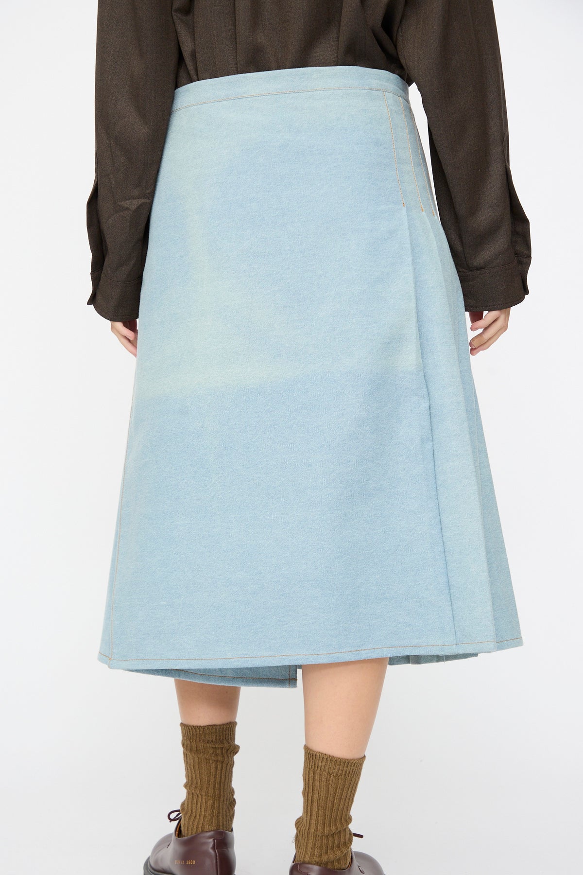 A Bless No. 52 Denimkilt in Denim Light Blue midi skirt worn with a dark brown long-sleeved shirt, olive green ribbed socks, and dark shoes, viewed from behind.