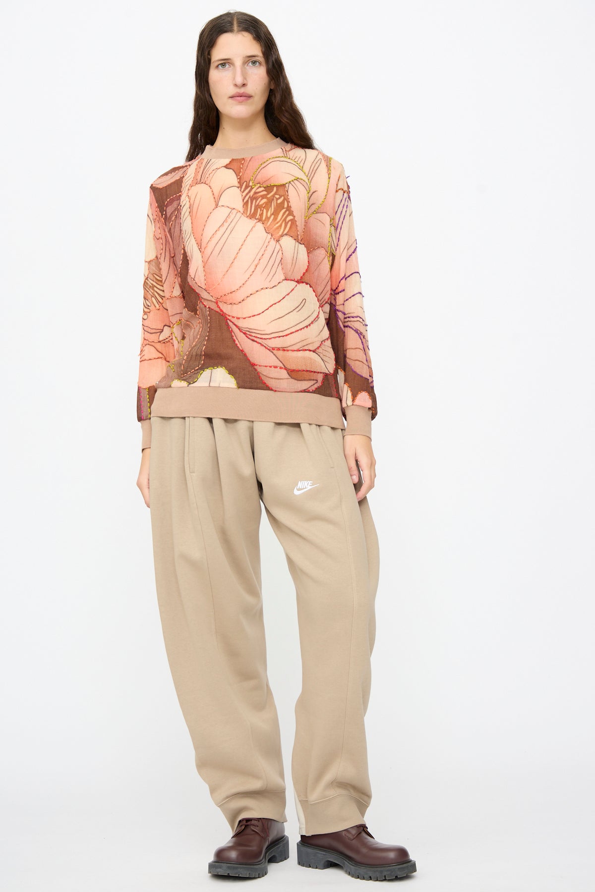 A person wearing a sheer, long-sleeved top with a large floral print, Bless No. 61 Overjoggingjeans in Beige and Sand, and dark brown shoes.