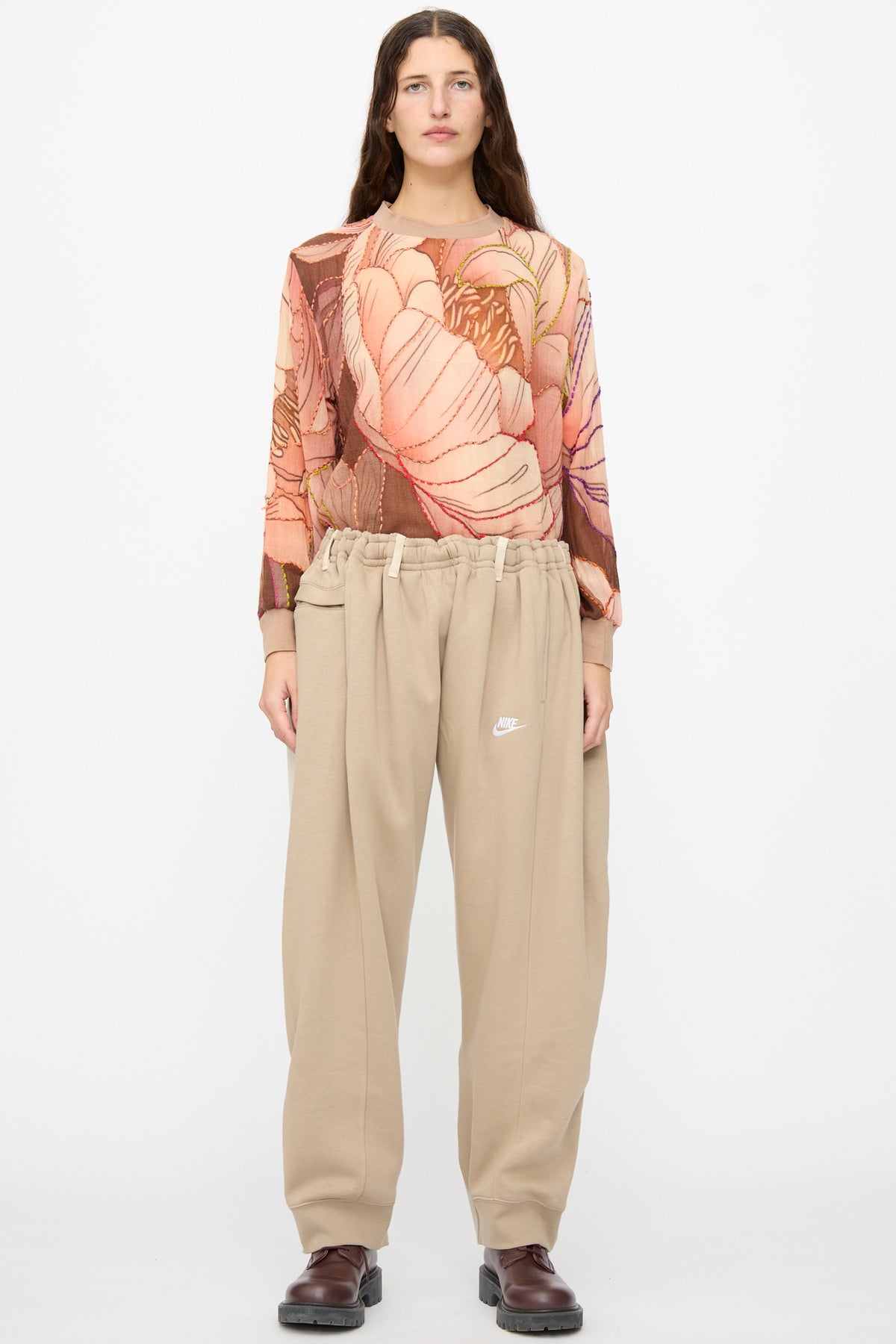 A person wears a sheer, long-sleeved floral top tucked into Bless No. 61 Overjoggingjeans in Beige and Sand paired with dark brown shoes.