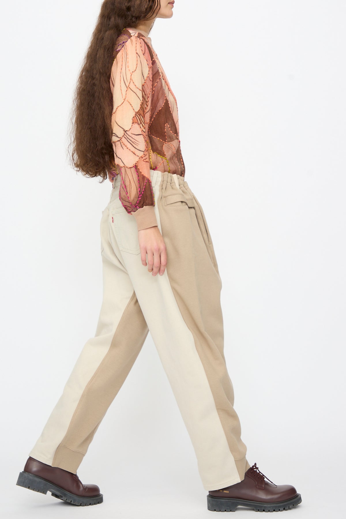A person walking in a patterned long-sleeve shirt and Bless No. 61 Overjoggingjeans in Beige and Sand, featuring a drawstring waist, elastic cuffs, and dark brown leather boots.