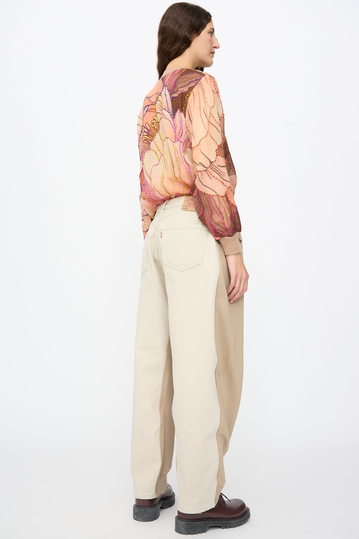 A person seen from behind wears a patterned, sheer long-sleeved top with a relaxed fit, Bless No. 61 Overjoggingjeans in Beige and Sand, and dark boots.