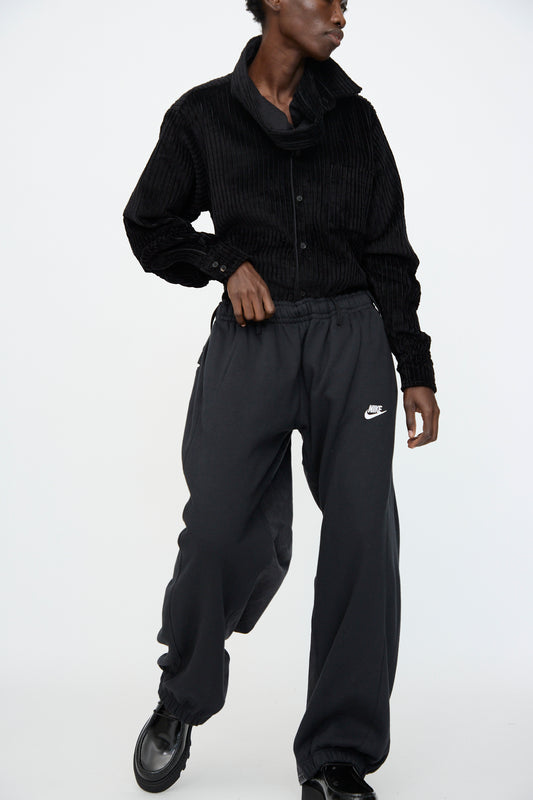 A person stands against a plain white background wearing a black corduroy shirt, Bless No. 61 Overjoggingjeans in Black with an oversized fit, and black shoes.